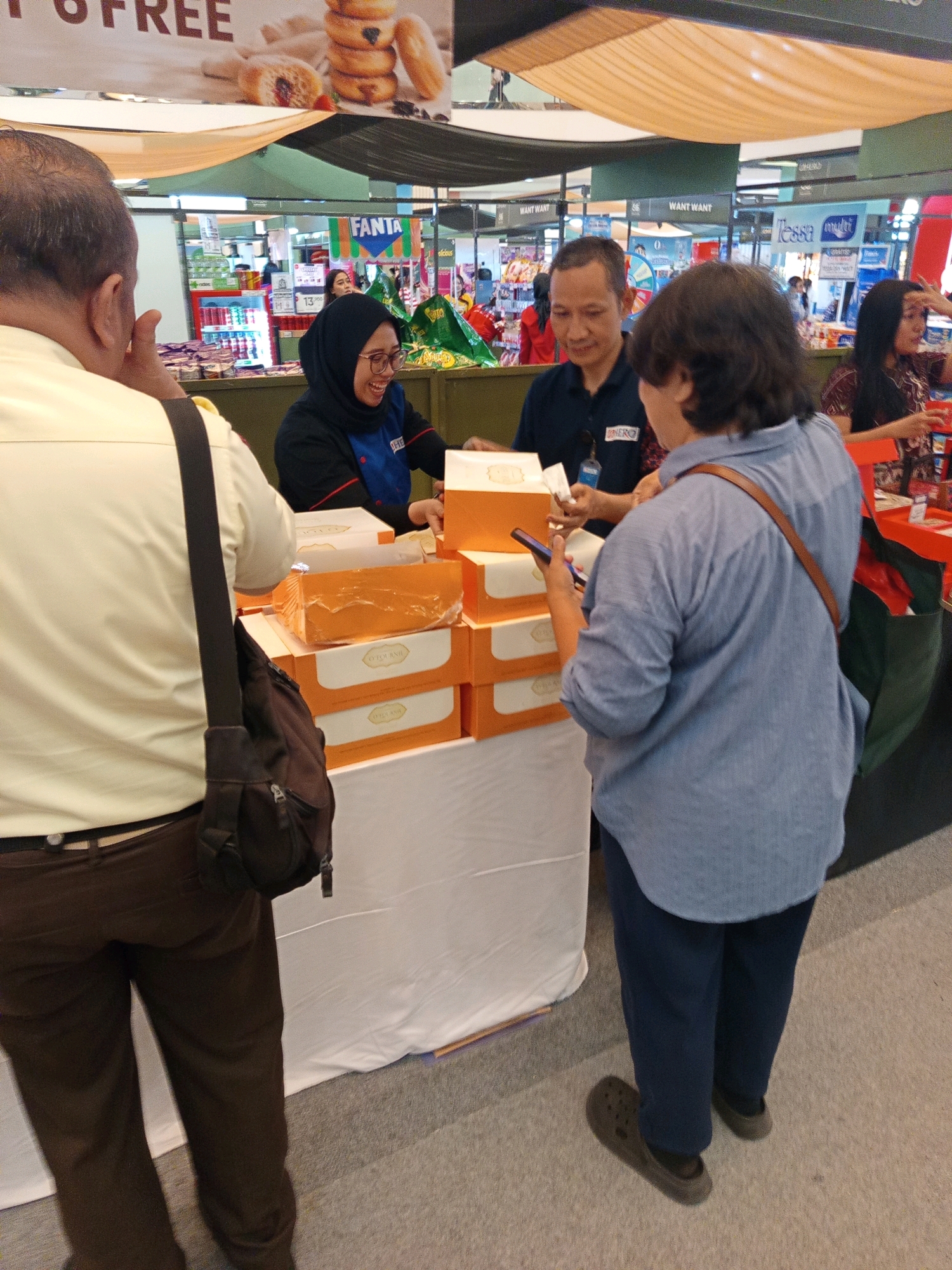 In West Jakarta, Indonesia, at what appears to be a supermarket or mall event, a group of individuals are gathered around a stall displaying boxes of pastries, possibly donuts, judging by the banner above which shows donuts and text indicating "FREE". A woman wearing a black hijab and a blue uniform with an embroidered logo is smiling and interacting with customers, holding a box. To her right, a man in a dark blue polo shirt, also with a logo, is engaged in a transaction, holding a white box. On the opposite side, a woman wearing a blue button-down shirt and dark pants, with a brown strap across her shoulder, is looking at her phone. Another person, viewed from the back and wearing a light yellow shirt and brown pants, has a dark bag slung over their shoulder. The stall is covered with a white cloth, and the background shows aisles of products in a retail environment with promotional banners. The lighting is bright and consistent with indoor retail spaces.