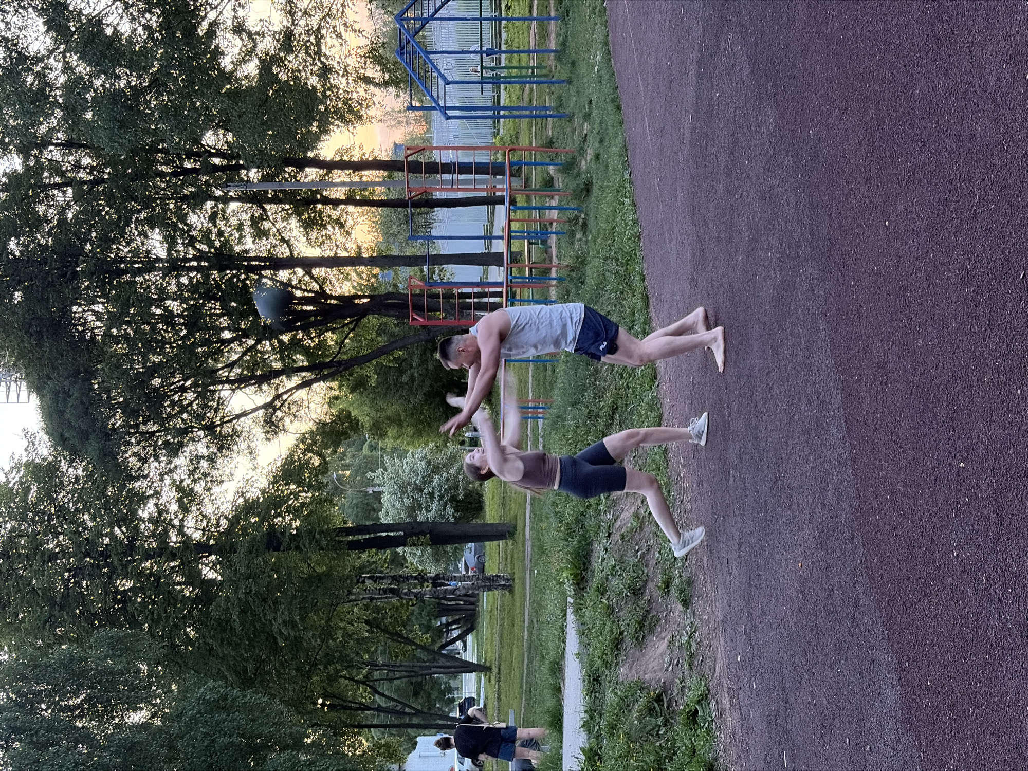 Two people stand opposite each other on a dark-colored track beside a grassy area, with trees and a playground visible in the background. A man wearing a gray tank top and dark shorts stands facing a woman wearing a brown tank top and dark shorts. Both people are extending their arms towards each other. Another person in dark clothing is doing push-ups at the bottom of the image. The location appears to be a public park in Izhevsk, Russia.