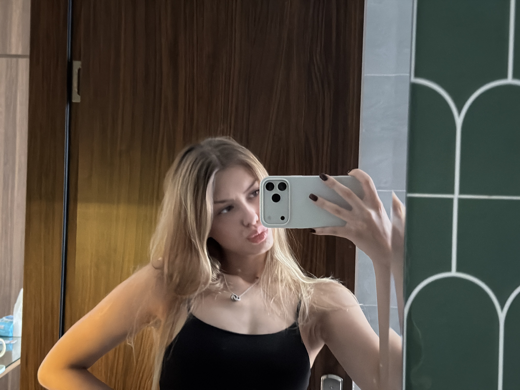 A young woman with blonde hair is taking a mirror selfie. She is wearing a black tank top and a silver necklace. She is holding a white smartphone in her right hand. Her left arm is bent at the elbow, with her hand resting on her hip. The background appears to be a room with wooden walls and a tiled wall with a green arch pattern. There is a doorway visible to the left, and a glimpse of a tissue box and other items on a shelf. The lighting suggests it is daytime. The woman has a pout on her lips, suggesting a playful or flirtatious mood. The image is likely taken in a bathroom or dressing room, possibly in a hotel due to the decor.