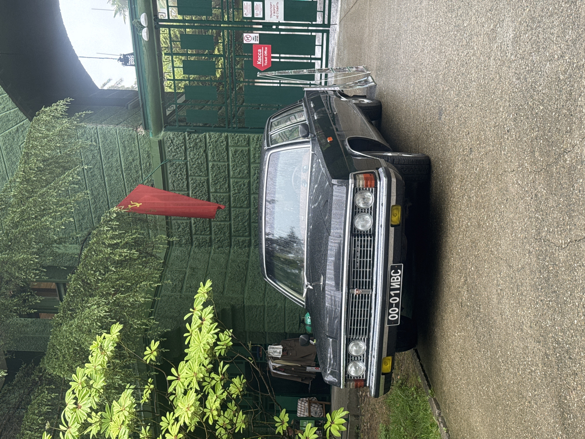 The image shows a dark-colored car, possibly a vintage model, parked in front of a green building with foliage. A red flag, likely a Soviet flag, is attached to the building. The car has a license plate that appears to be Russian, and it is parked on a paved surface. In the background, there's a green metal gate with signs, including one indicating "Kassa" (Ticket Office). The scene is in Sochi, Russia.
