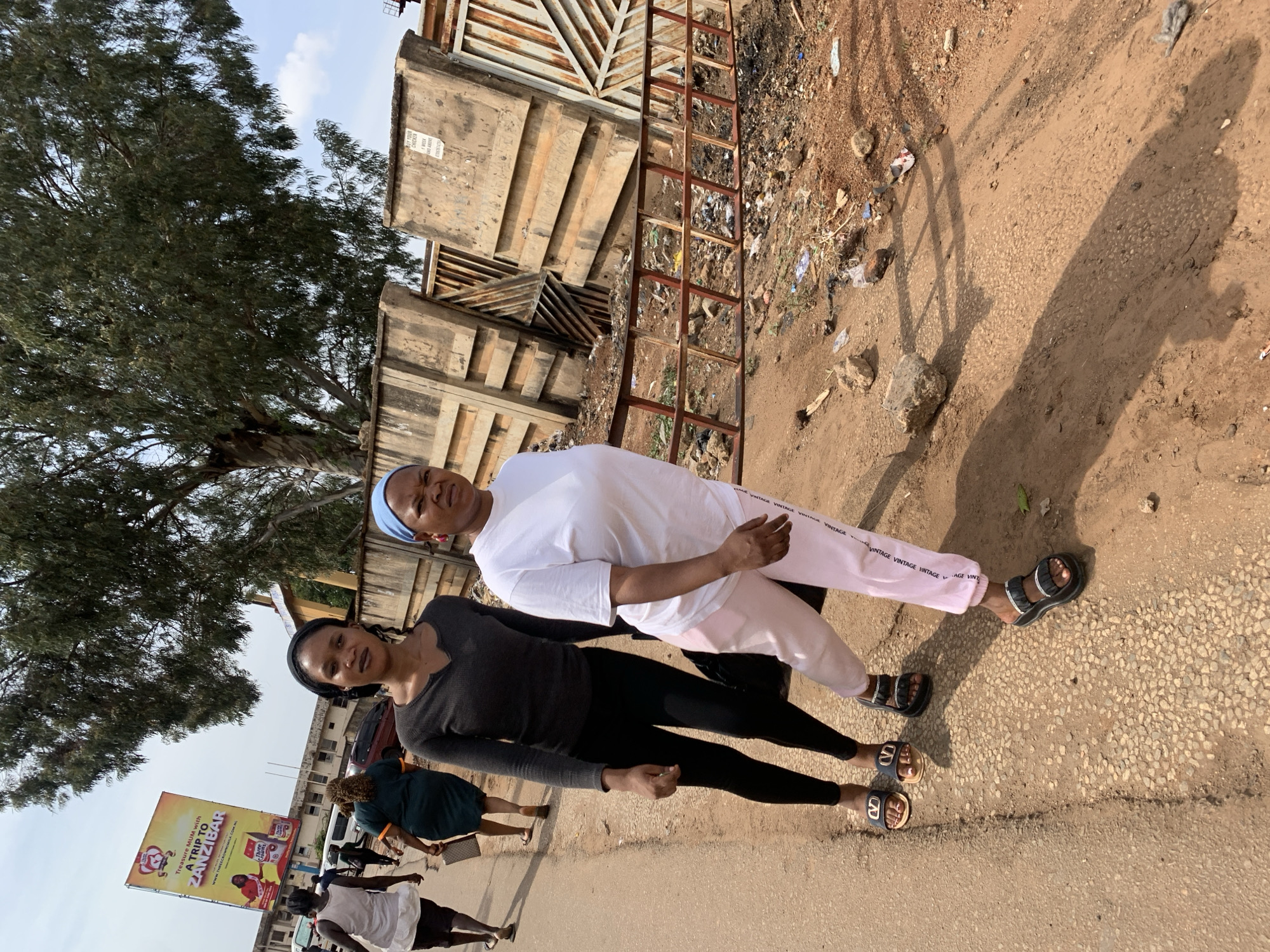Two women are prominently featured standing side-by-side on a sunlit dirt and gravel path with patches of asphalt, likely a street in Jos, Nigeria. The woman on the left (viewer's left) wears a black long-sleeved top, black leggings, a dark headwrap, and open-toed sandals with a distinctive "V" logo. She looks towards the camera with a slight smile. The woman on the right wears a white t-shirt, light pink sweatpants with "VINTAGE" repeatedly printed on them, a light blue headwrap, and black platform sandals. She gazes slightly past the camera with a neutral expression.
The scene is outdoors under bright daylight, indicated by the clear sky and long shadows cast on the ground by the subjects and a metal ladder-like object. This suggests either morning or late afternoon. The ground is uneven, dotted with small rocks and some scattered litter.
In the background, a large tree with dense green foliage occupies the upper left portion of the image. Further down the street, several other individuals are visible walking, including one person in a green dress and another in a white outfit. A large yellow billboard stands out on the left side of the street, advertising "Treasure MUM with A TRIP TO ZANZIBAR," along with the text "REECROWNSM" and a partially visible website address "www.MECROWNSM.COM.NG".
To the right, behind the two main subjects, a tall, rusty metal gate or fence structure is visible, backed by concrete walls. On one of these concrete walls, a sign partially reads: "PICK YOUR CHICKEN," "FREE," "1 WEEK," "AND ABOVE," and "DELIVERY."