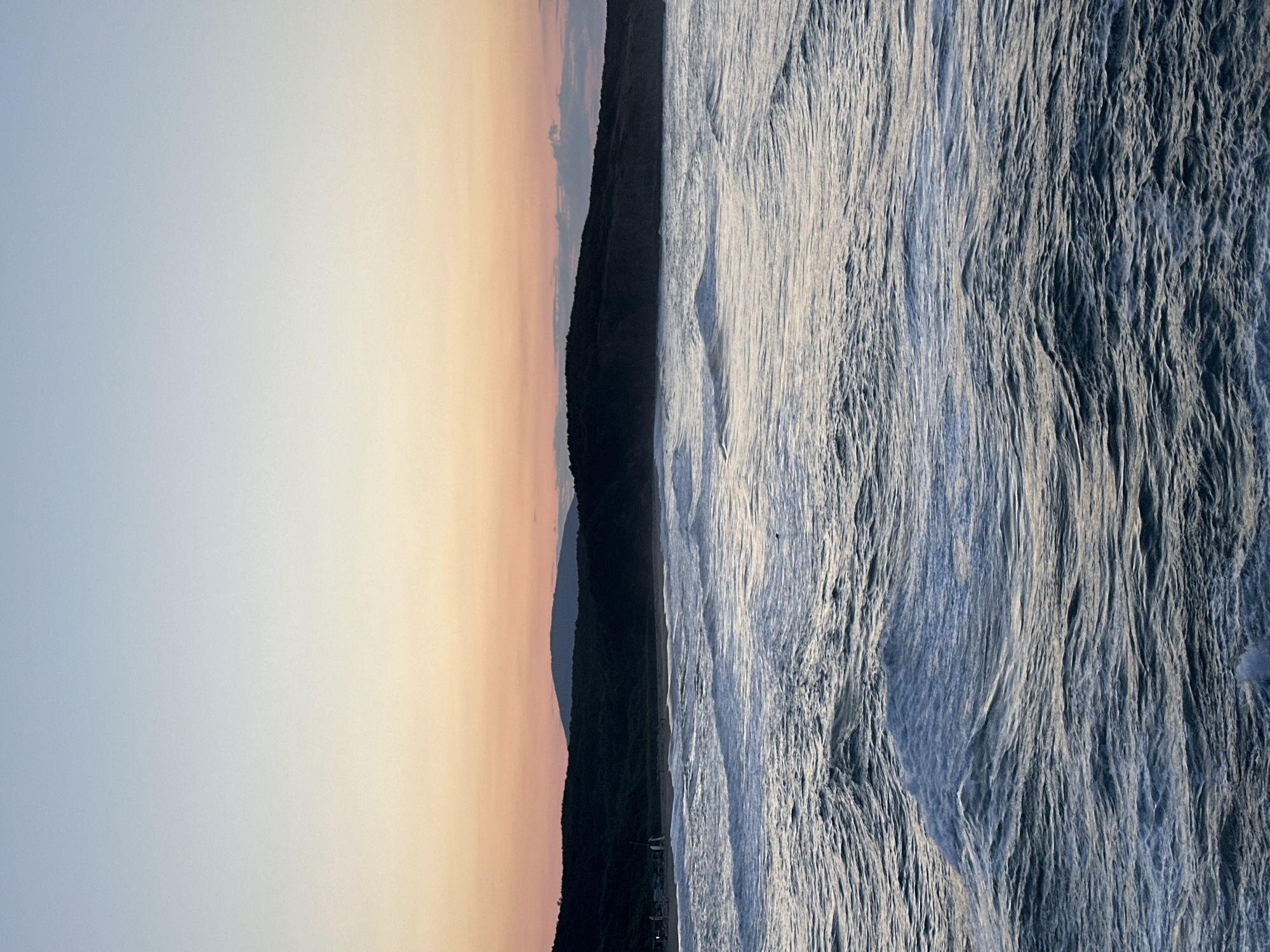 The image depicts a view of the Black Sea near Sinemorets, Bulgaria, during a sunset. The sky transitions from a light blue at the top to a warm orange and pink hue near the horizon. A dark, tree-covered hill forms the coastline, with a distant mountain range visible beyond it. The foreground is dominated by choppy, greyish-blue waves of the sea, showing white foam and crests. A few small figures are visible on the beach in the distance, and a single bird is silhouetted against the water. The overall atmosphere is tranquil and serene, characteristic of a coastal sunset.
