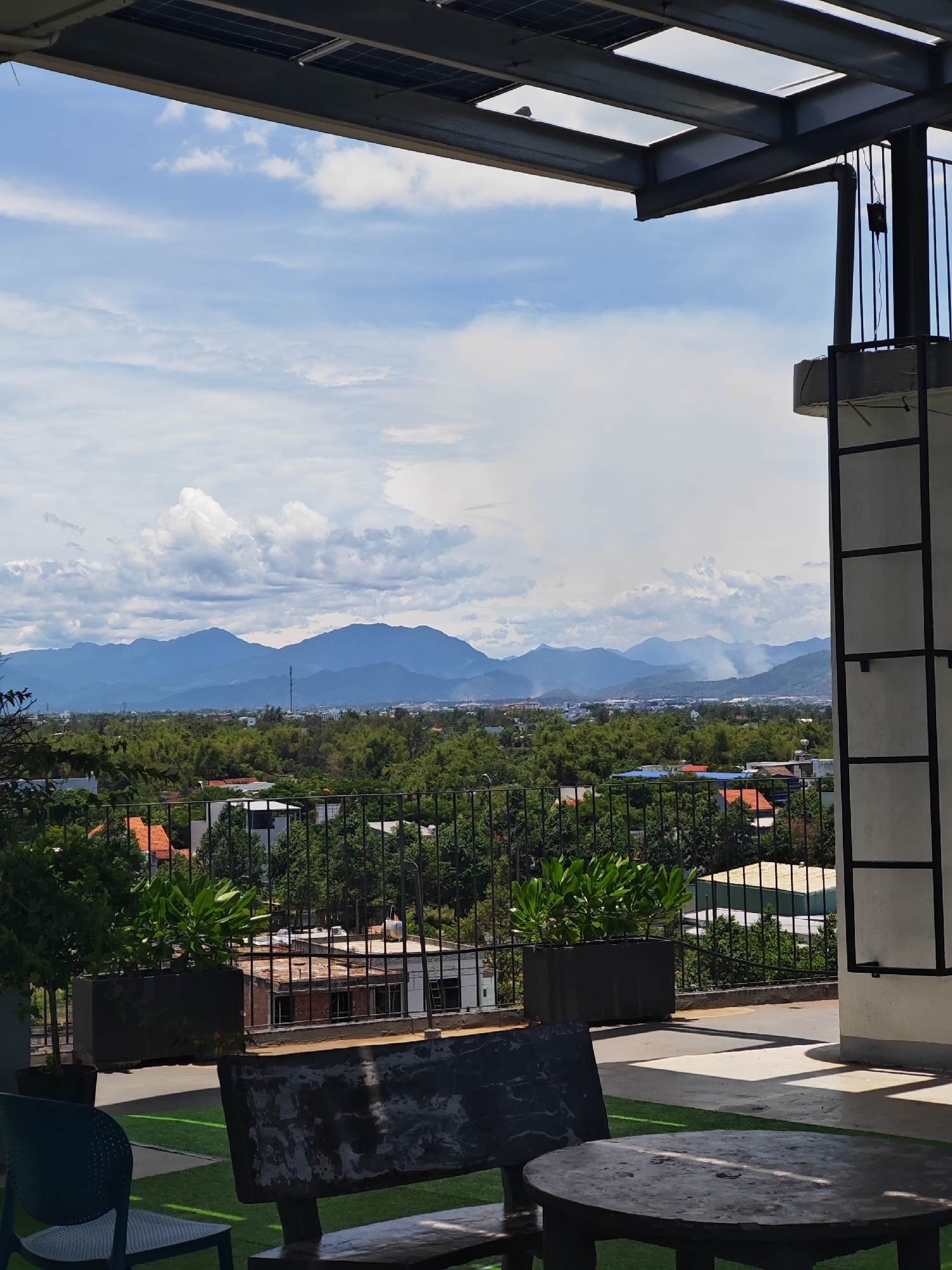 The image is a landscape view from a high-rise balcony in Da Nang, Vietnam, offering a panoramic vista of the city and surrounding mountains under a partly cloudy sky. In the foreground, a dark-colored bench and a round table are set on a green, artificial turf area, suggesting an outdoor seating or relaxation space. To the left, a dark blue chair is partially visible. Beyond the seating area, a black metal railing encloses the balcony, behind which planters with green foliage are arranged. The middle ground of the image features a dense expanse of green trees interspersed with houses and buildings, their roofs varying in color and material. Further in the distance, a mountain range stretches across the horizon, its peaks shrouded in a soft, hazy blue due to atmospheric conditions. Wisps of smoke are visible rising from pockets within the foothills of the mountains, indicating possible agricultural burning or industrial activity. The sky above is a mix of bright blue and scattered white clouds, with some larger, more dramatic cloud formations visible, particularly towards the left side of the frame. The upper portion of the image shows a section of a modern building's roof structure, including what appears to be solar panels and exposed metal framework, hinting at the urban environment from which the photograph was taken.