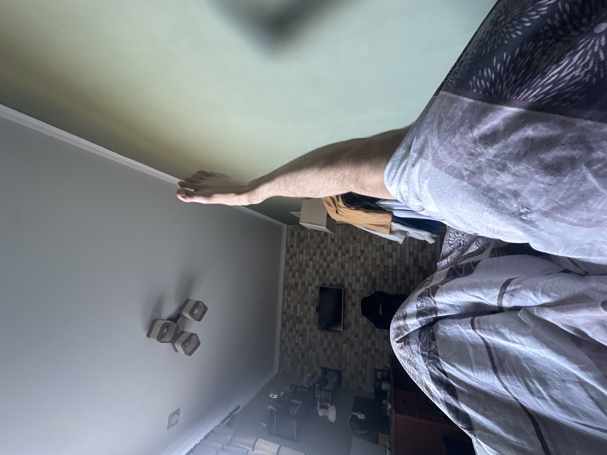 A foot and leg extend into a room with a bed, clothes, and furniture. The ceiling has a modern light fixture. A television is mounted on a brick-patterned wall. The location is Unknown, Unknown.