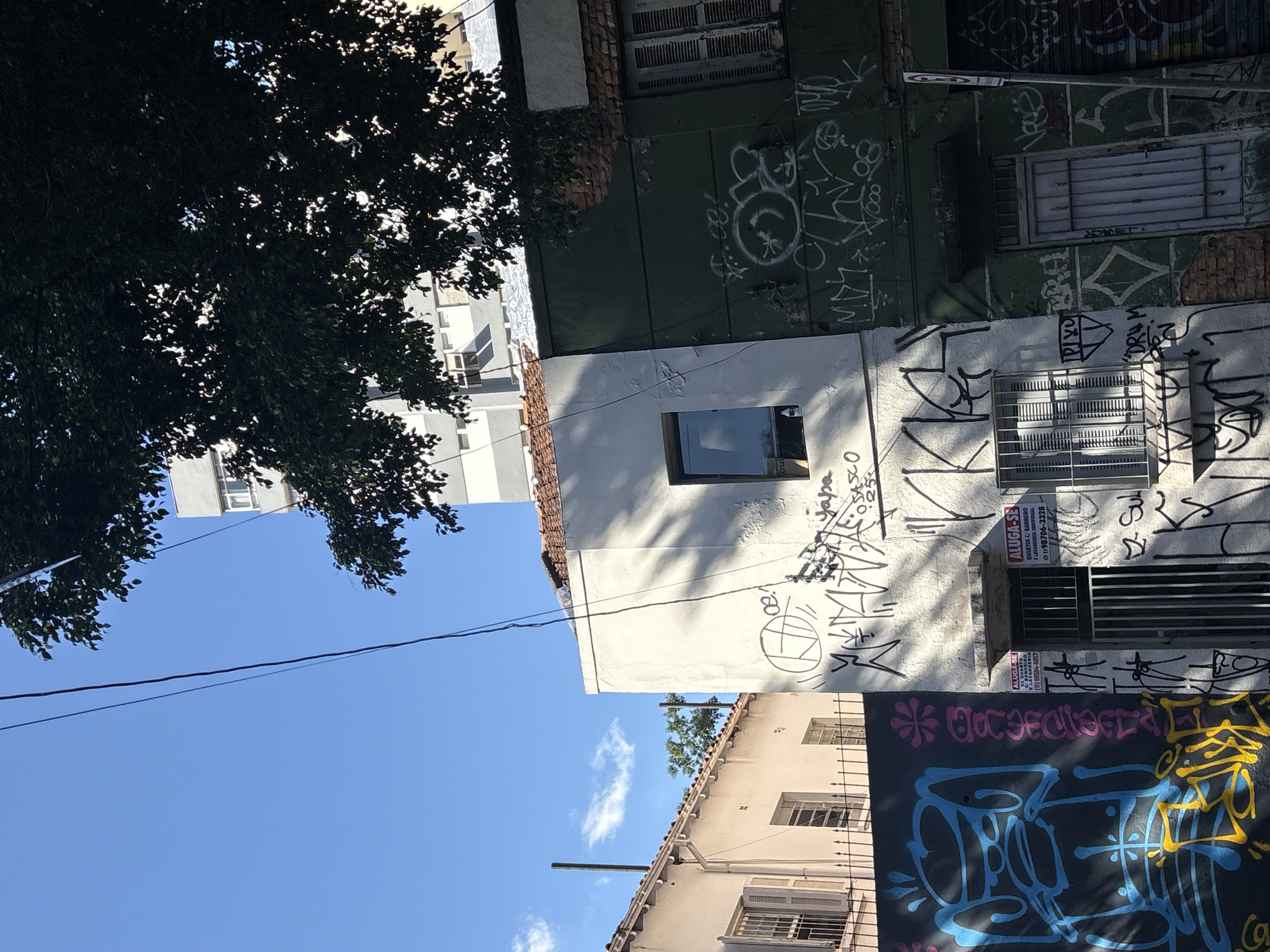 The image shows an exterior view of a building in the city of São Paulo, Brazil, on a sunny day. The building has a white facade covered in graffiti and a green facade, also with graffiti. There are several windows, some with shutters, and a metal door. Above the building, a tree casts shadows, and power lines are visible against a clear blue sky. Two signs advertising rooms for rent are also visible. One sign states "ALUGA-SE QUARTOS C/ BANHEIRO E COZINHA INDIVIDUAL" with a phone number. The overall scene depicts an urban environment with signs of decay and artistic expression through graffiti.