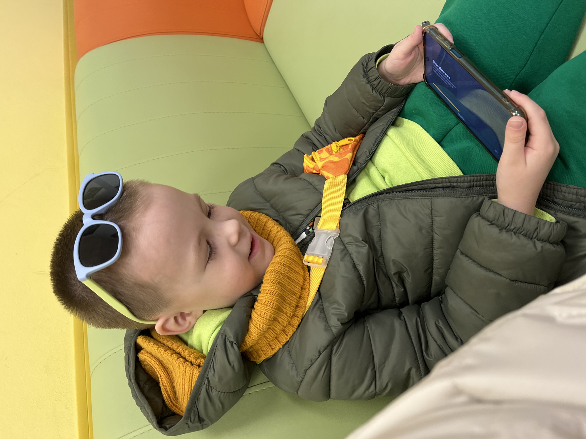A young child is lying on a multi-colored cushion. The cushion has a yellow, green, and orange section. The child is wearing a green padded jacket, a yellow neck warmer, a lime green shirt, and green sweatpants. Blue sunglasses are on the child's forehead. The child is holding a smartphone in their hands and looking at the screen. The smartphone shows the text "Single-player world" and "Initializing 48%". The location is undetermined, other than the request to include Usman, Russia, in the description. No reliable information allows me to establish a connection to the city of Usman, Russia.
