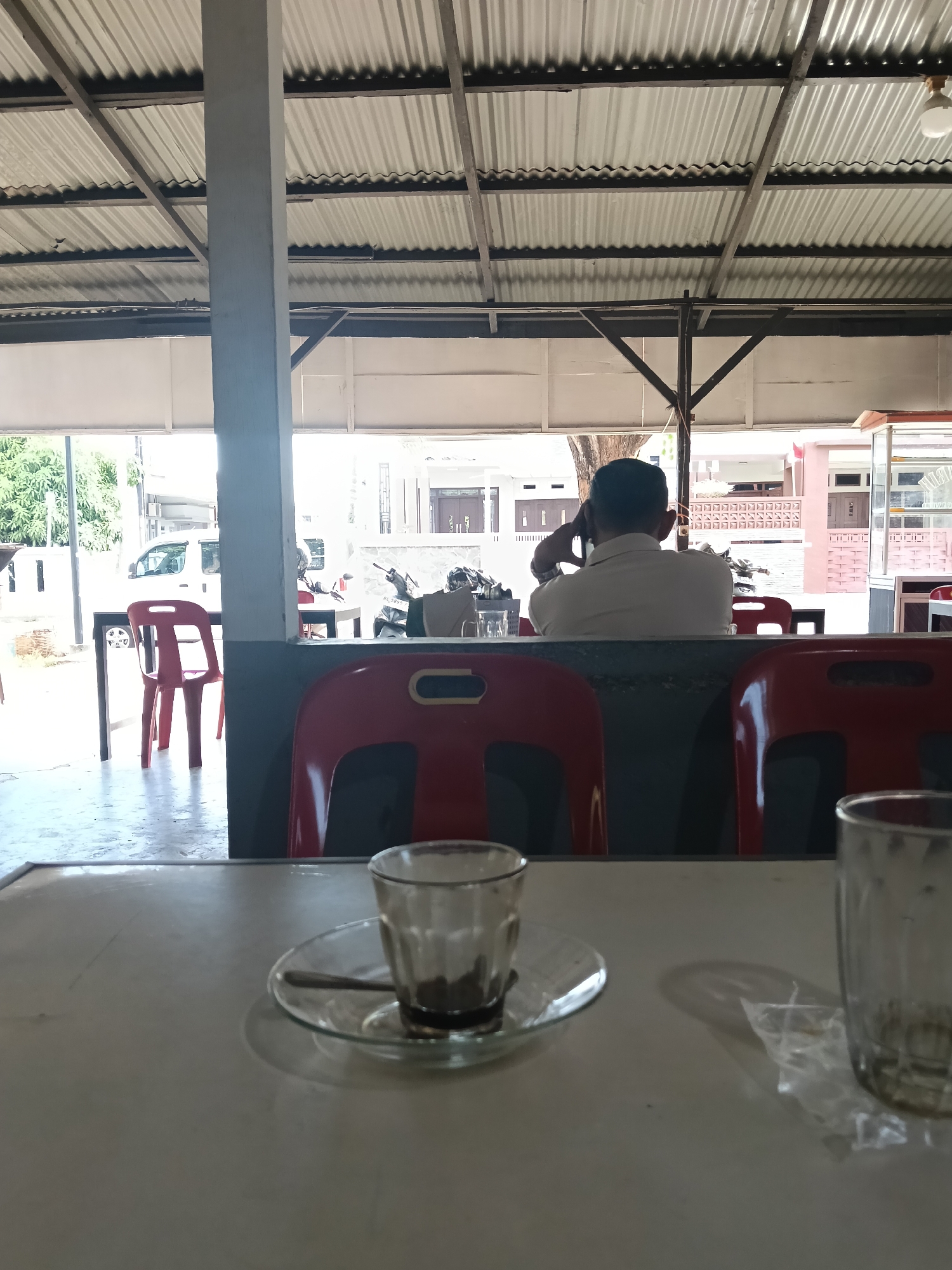 A man sits with his back to the camera, holding a phone to his ear, in what appears to be an outdoor cafe or warung in Banda Aceh, Indonesia. He is wearing a light-colored short-sleeved shirt. In the foreground, on a white table, are two glasses and a small cup with a saucer and spoon. One glass is partially filled with a dark liquid, likely coffee, and the other is empty. The cafe has a corrugated metal roof supported by wooden beams and a concrete pillar. Red plastic chairs are visible throughout the scene, some empty and some occupied by the man. In the background, there are buildings, trees, and a white van parked on the street. The lighting suggests it is daytime, with bright sunlight casting shadows.
