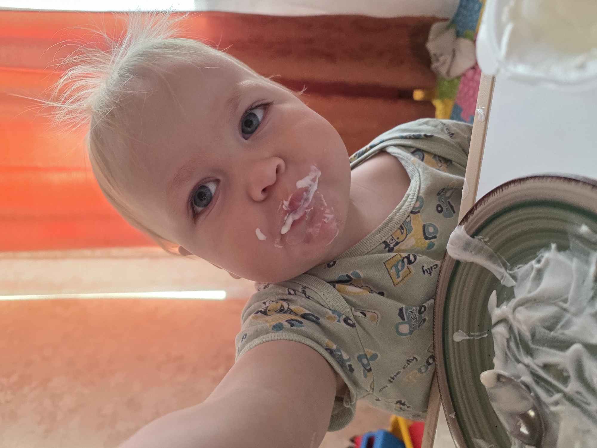 A close-up shot captures a baby with fair skin and light blond hair. The baby's face is partially covered with white food residue around the mouth. The baby is wearing a light green onesie patterned with vehicles and letters. A bowl of white, creamy food is partially visible. The location is presumed to be in a domestic setting in the city of Chaikovsky, Russia, inferred from the query.