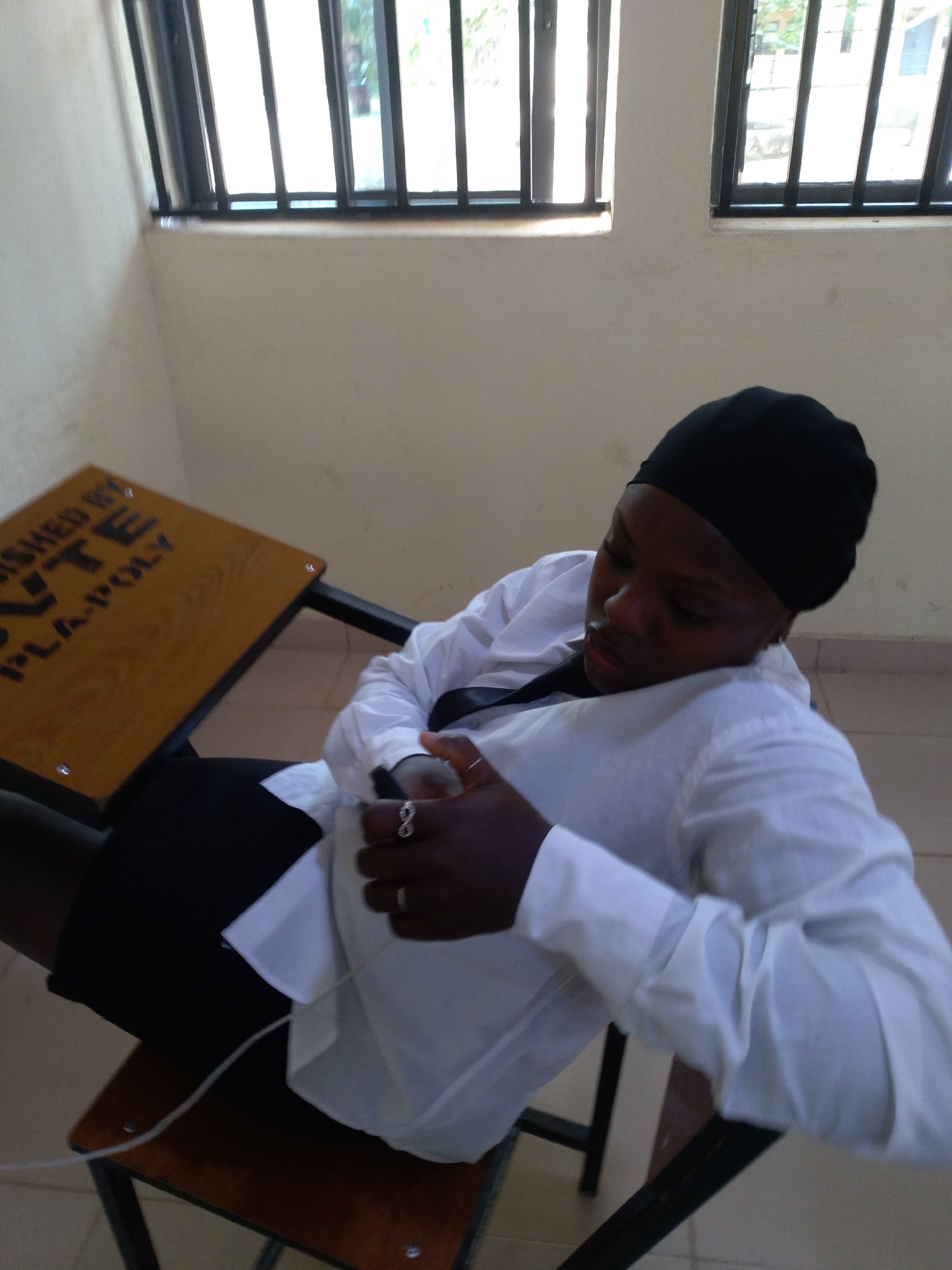 This image captures an indoor scene in Jos, Plateau State, Nigeria, depicting one individual seated in what appears to be a classroom or study area. The location is identified by the inscription "PLA-POLY" visible on the desk, referring to Plateau State Polytechnic, which is situated in Jos, Nigeria.
The subject is a person, likely female, with dark skin, wearing a black head covering, a white long-sleeved button-down shirt, and dark-colored lower garments. She is seated, leaning back in a chair, with her head tilted downwards, looking at her hands. A thin white cable extends from her hands, running down towards the chair's seat, suggesting she is interacting with or charging an electronic device, though the device itself is not clearly visible. She wears two rings on her right hand: one on the ring finger and another, with an infinity symbol design, on the middle finger.
The environment features light-colored (off-white) walls, which show some minor marks or discoloration. In the background, two windows with black metal security bars are visible, allowing natural light into the room and offering a glimpse of greenery outside. The floor is tiled with light-colored squares. A wooden-topped desk with a black metal frame is positioned to the left of the person. The visible inscription on the desk reads, "ISHED BY R.O.N.T.E. PLA-POLY." The chair the person is using has a wooden seat and a black metal frame. The overall atmosphere suggests a functional, naturally lit institutional setting.