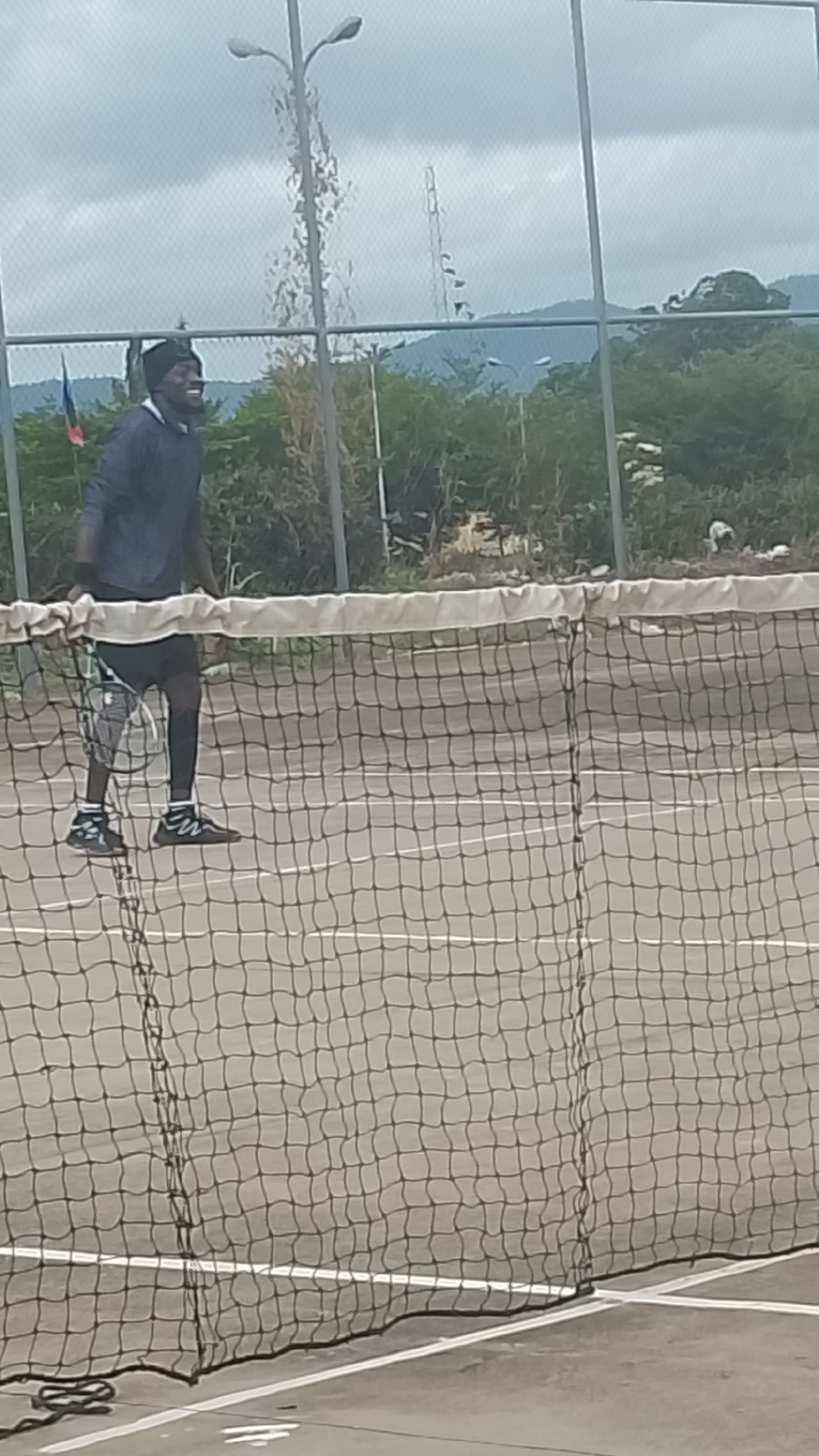 The image depicts an outdoor tennis court in Lokoja, Nigeria, under an overcast sky. A single individual, appearing male, is positioned on the left side of the court, behind the net. He wears a dark beanie, a dark long-sleeved top, dark shorts over dark leggings, and dark athletic shoes with white soles. He is holding a tennis racket with his left hand, angled downwards, and exhibits a smiling facial expression.
The tennis net, featuring a white top band, extends across the lower half of the frame. The court surface is light grey with visible white boundary lines. Behind the player and the net, a chain-link fence is present. Beyond the fence, dense green vegetation is visible. In the background, a small flag displaying red, blue, and black colors is partially seen on the left. Several streetlights with dual lamps are scattered across the midground and background, and a tall lattice-style communication tower is visible on the right. Distant mountains or hills are silhouetted against the grey, cloudy sky.