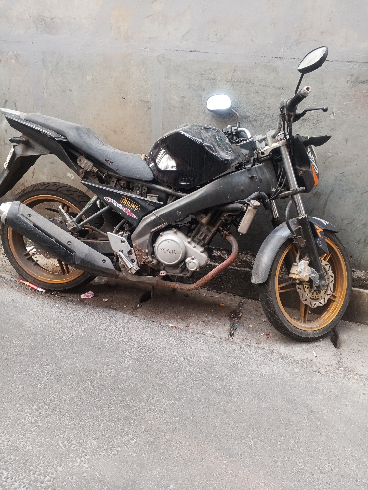 A weathered black Yamaha motorcycle is parked against a grey, concrete wall on a city street in West Jakarta, Indonesia. The motorcycle appears to be in a state of disrepair, with a dirty fuel tank covered in a plastic wrap, a worn seat showing signs of damage, and various parts appearing rusted or dirty. The exhaust pipe is also rusted. The front tire and rim are gold in color, as is the rear rim. The street itself is paved and appears to have some debris scattered on it. The scene is lit by natural daylight.