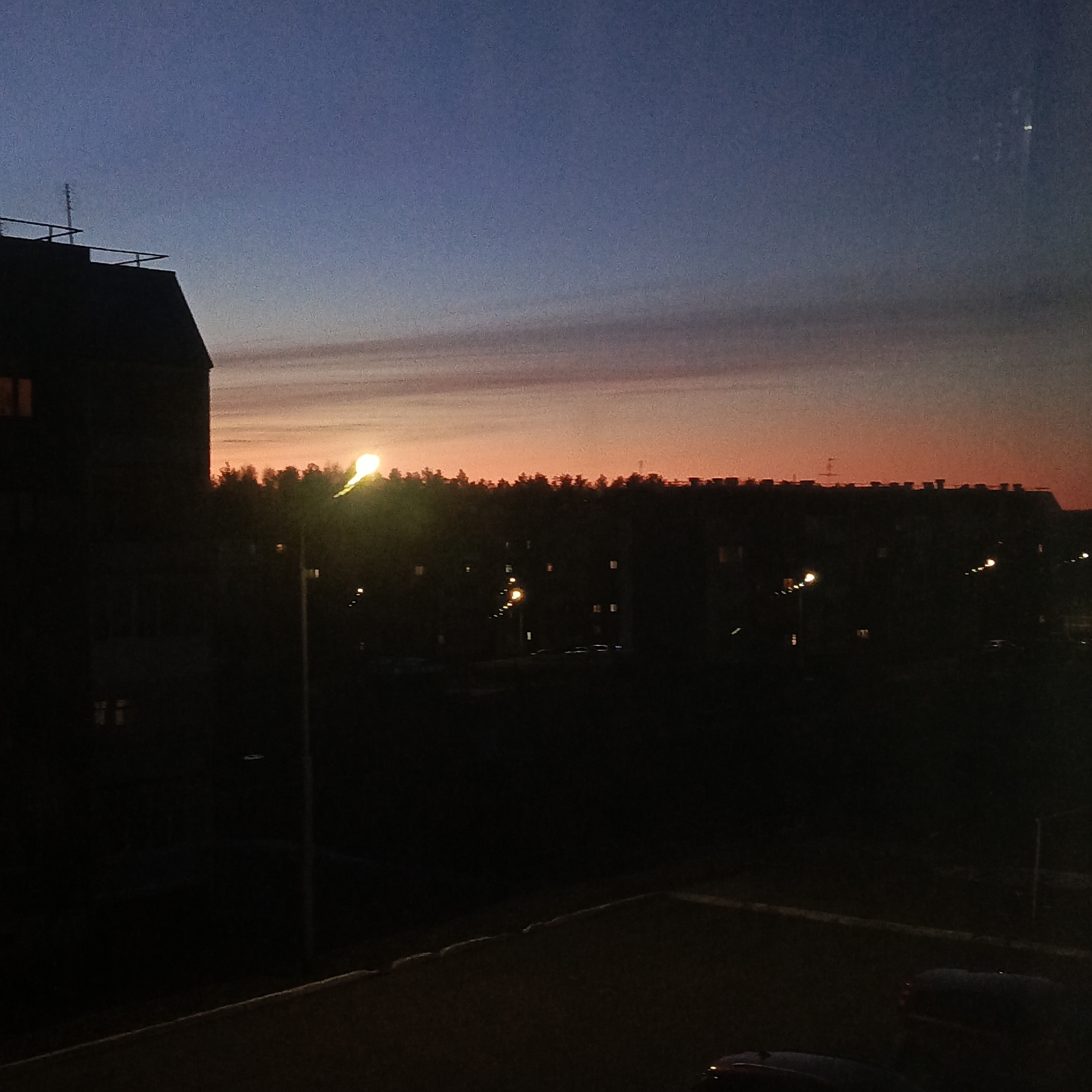 The image depicts a twilight scene in Марковский, Russia. The sky transitions from a deep blue at the top to orange on the horizon, partially obscured by a row of dark trees and buildings. Several street lights illuminate the area, one prominently in the foreground. A large building is visible on the left, with an antenna on its roof. The foreground is obscured in darkness, partially revealing a paved area and the top of parked cars.
