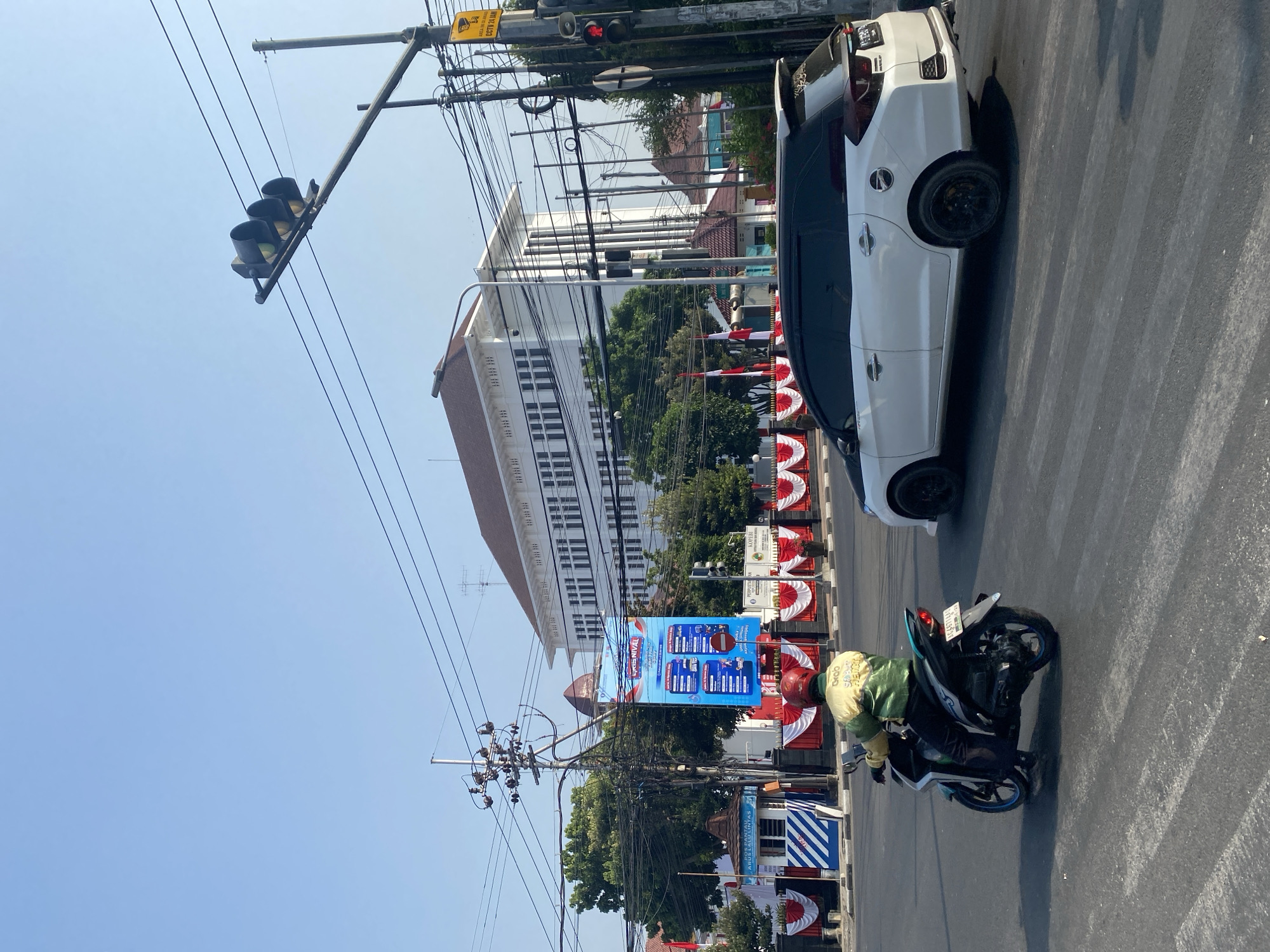 The image captures a street scene in Kediri City, Indonesia, characterized by clear blue skies and bright sunlight. In the foreground, a motorcyclist wearing a green and yellow jacket with "Grab" branding rides through a crosswalk. Behind the motorcycle, a white SUV with tinted windows is stopped at an intersection.
Dominating the mid-ground is a large, white, multi-story building with a distinct architectural style featuring numerous windows and decorative elements. In front of this building, a banner hangs vertically, displaying various images and text in blue and red. The building is flanked by trees and a festive display of red and white draped fabric, suggesting a celebration or event.
The background is filled with a complex network of electrical wires and poles, indicative of urban infrastructure. A traffic light hangs overhead, displaying yellow signals, and a traffic camera is mounted on a pole with a yellow sign. The overall environment appears to be a busy urban intersection.