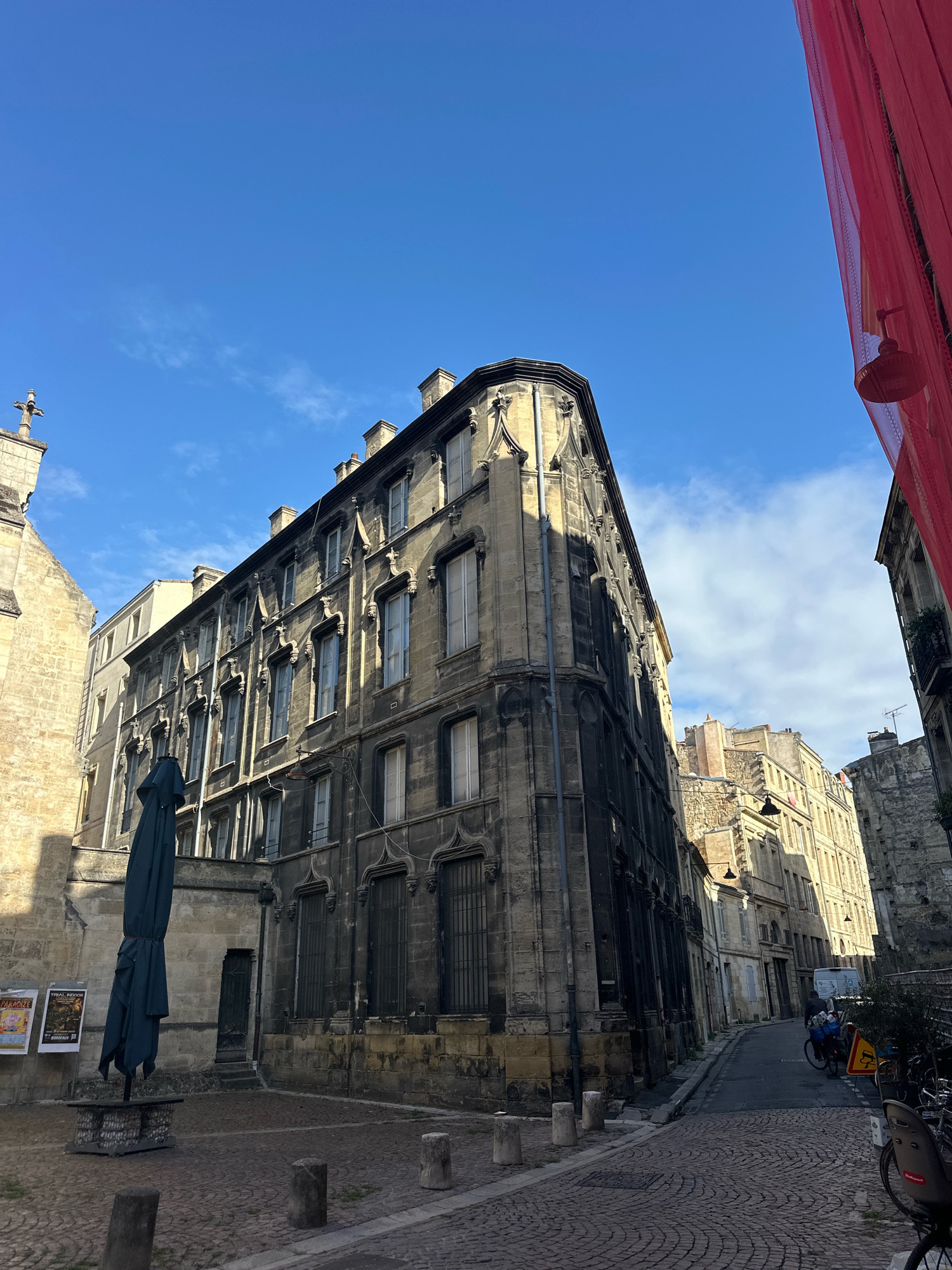 The image shows a street scene in Bordeaux, France, on a bright, clear day with scattered clouds. The architecture suggests an old European city, likely with historic buildings. In the foreground, a cobblestone street curves to the right, lined with stone buildings.
Dominating the left side of the image is a tall, dark, ornate building with multiple stories, featuring numerous windows and intricate stone carvings. Adjacent to it on the left is a lighter-colored stone building, possibly part of a church or another historical structure, with a cross visible at its peak. In front of the dark building, a large, dark green umbrella is closed and leaning against a low stone structure. Posters are affixed to the wall of the lighter building to the left of the umbrella.
The street recedes into the background, showing more buildings with similar architectural styles, some appearing older and more weathered than others. A white van is parked on the right side of the street, and several bicycles are visible, some parked and others with riders. A person is seen on a bicycle on the right side of the street, moving away from the camera.
The right side of the image features a vibrant red fabric draped vertically, obscuring part of the view into a narrow alleyway or another street. The sunlight casts strong shadows, indicating the time of day. The overall impression is a picturesque urban setting with a blend of historic architecture and everyday street life.