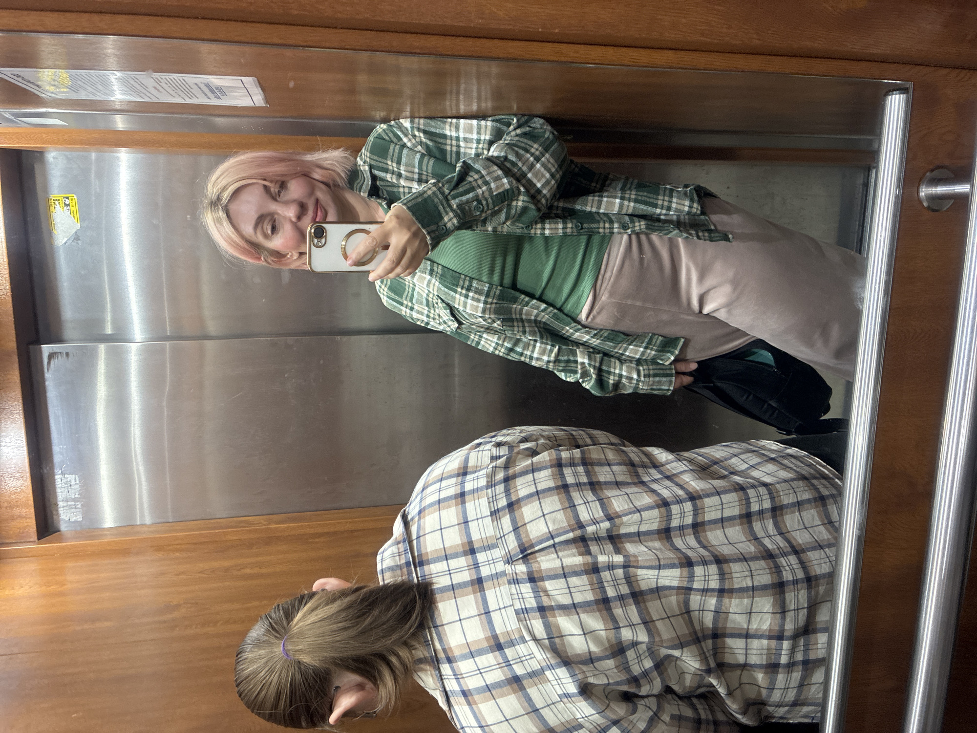 The image is a mirror selfie taken inside an elevator or a room with mirrored walls, rotated 90 degrees counter-clockwise.
In the reflection, a woman with short, light pink hair smiles softly while holding a white smartphone with a gold ring holder to take the picture. She is dressed in a green and white plaid flannel shirt over a green ribbed top and light brown pants. A black bag is partially visible near her leg.
In the foreground, seen from behind, is another person with brown hair tied back in a ponytail with a purple elastic. This individual wears a white, blue, and brown plaid flannel shirt.
The setting features wooden paneling on the side walls and ceiling, complemented by stainless steel panels on what appears to be an elevator door. Silver-colored metal handrails or trim are visible on the right side of the mirror.
On the stainless steel panel reflected in the mirror, there are two pieces of paper: a white rectangular sheet with small, illegible black text, and a yellow sticker, also with small, illegible black text, possibly a warning or informational sign. The scene depicts a casual moment, likely in Sochi, Russia.