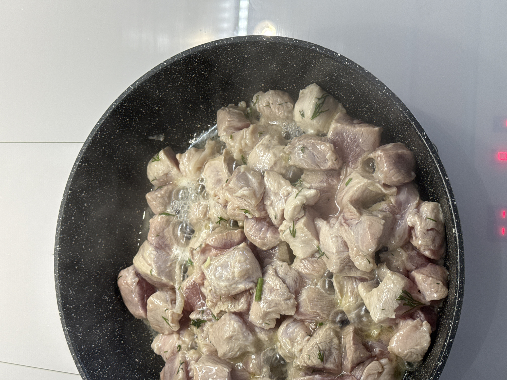 A high-angle, close-up shot shows a black frying pan filled with pale, raw meat cubes. The meat is partially submerged in a light-colored liquid, and some dill sprigs are visible among the cubes. Steam rises from the pan. The pan sits on a white surface, likely a stovetop. Red indicator lights are visible on the right side of the image. The location is potentially in the city of Лобаново, Russia.