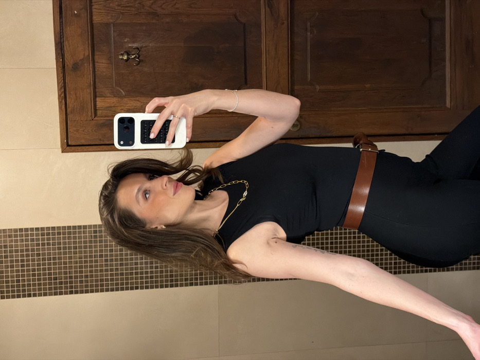 An adult female is depicted from the mid-torso up, taking a mirror selfie indoors. She has long, dark brown hair and wears a black sleeveless top, black trousers, and a wide brown leather belt with a silver-toned buckle. Accessories include small gold-colored hoop earrings, a gold-colored chain necklace with white elliptical accents, a ring on her left ring finger, and a delicate bracelet on her left wrist. She holds a white smartphone with a black textured grip in her left hand, angled towards her reflection, and is looking towards her right, away from the camera, with a slight smile. The background consists of a tiled wall on the left, featuring light beige rectangular tiles alongside a vertical strip of smaller brown square mosaic tiles. To the right, a dark brown wooden door with visible grain and a small metallic hook or knob is present. The specific location within Morinj, Montenegro, cannot be definitively ascertained from the visual evidence provided.