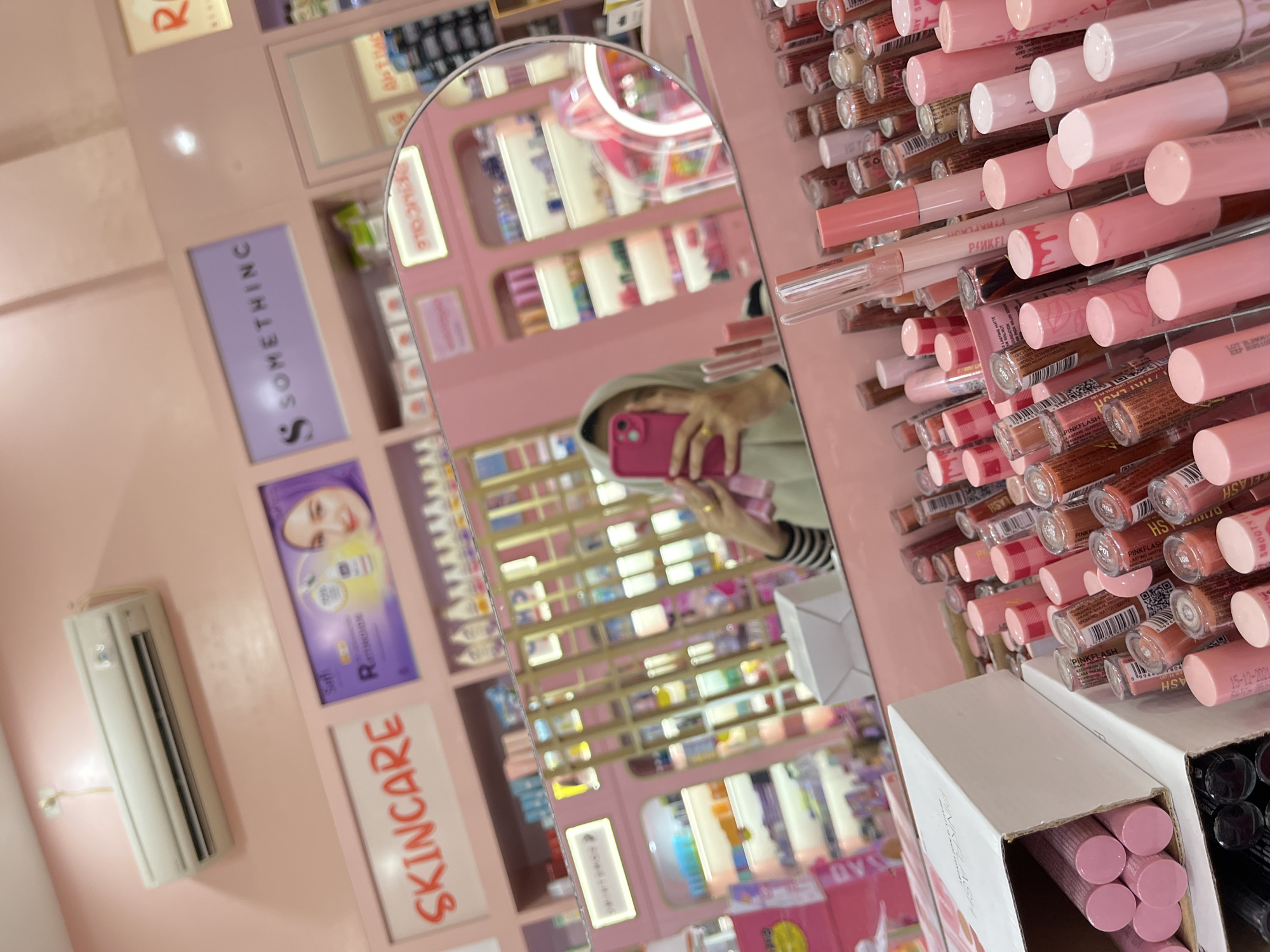 The image depicts the interior of a brightly lit cosmetics store, predominantly decorated in shades of pink. In the foreground, an assortment of lip glosses and lipsticks are neatly arranged, with some in open boxes. A mirror reflects the scene, showing a person holding a pink smartphone and taking a selfie. Behind the display of lip products, shelves stocked with various beauty items are visible. Prominently displayed on the wall are signs that read "SKINCARE" and "S SOMETHING." An air conditioning unit is mounted high on the pink wall. Based on available information, the store is likely located in Lhokseumawe, Indonesia, given the presence of the "S Something" brand which has a strong presence in the region. The overall atmosphere is that of a clean and well-organized retail environment focused on beauty products.