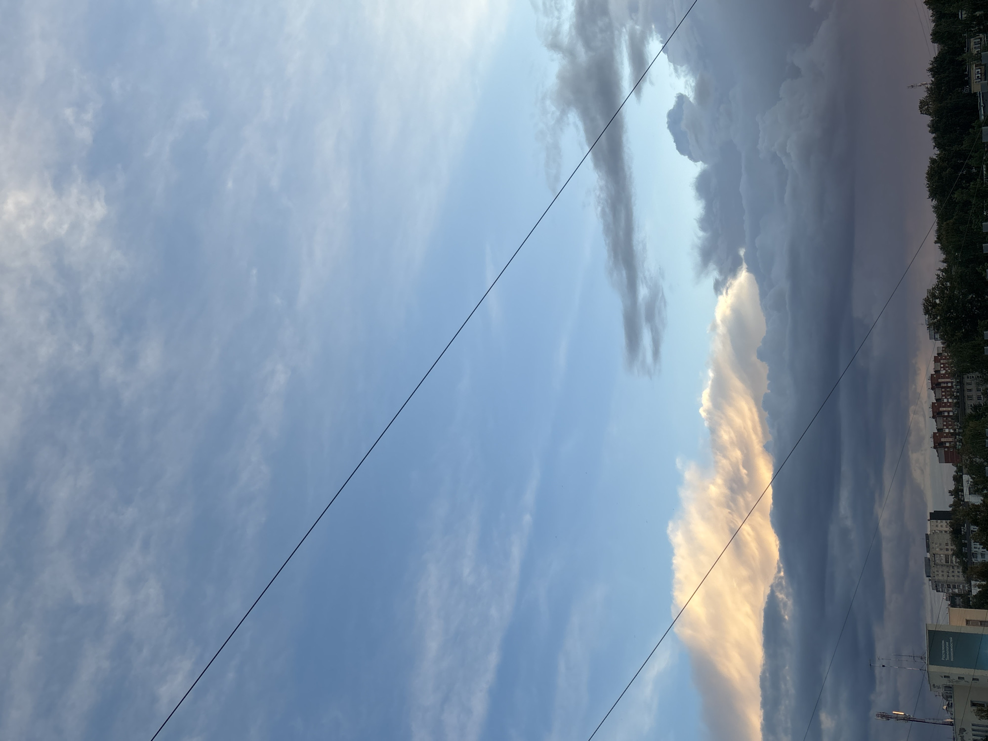 The image depicts a sky with a mix of light blue and dark storm clouds. Two electrical wires stretch diagonally across the frame. The lower right corner shows buildings and trees, suggesting an urban environment in the city of Chaikovsky, Russia. No people are visible.