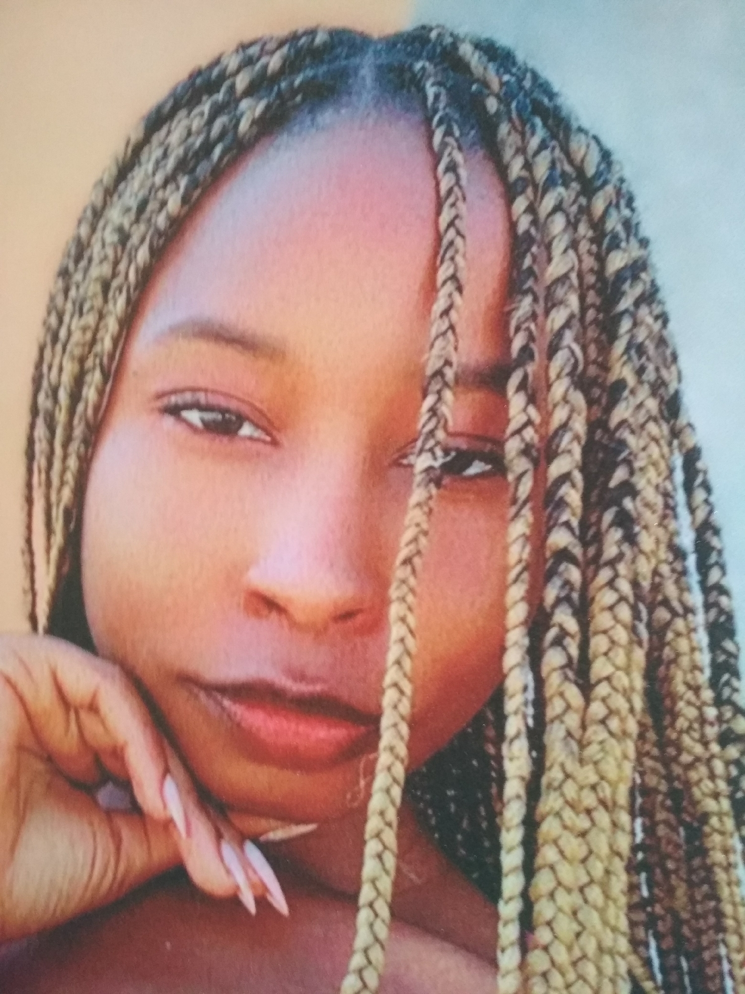 A close-up portrait of a young woman with dark skin and braided hair. Some of the braids are blonde and black. She is resting her face on her hand, her fingers are long with sharp nails. The background is blurred. Location is in the city of Unknown, Unknown.