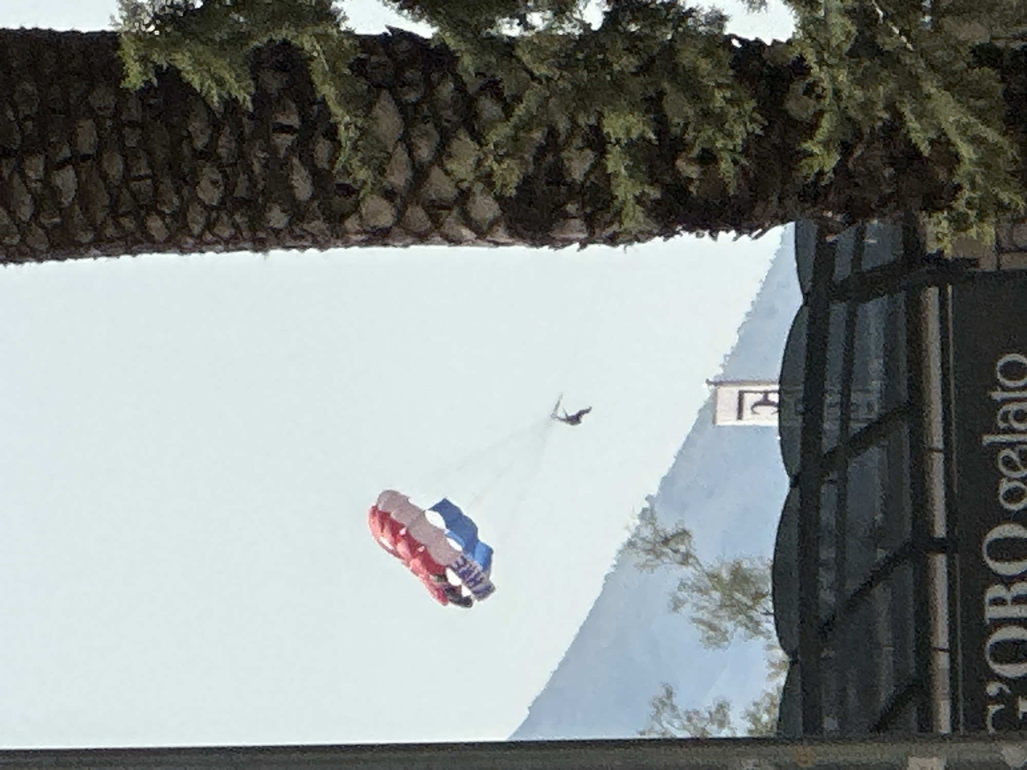 A person is parasailing through a bright, hazy sky over Budva, Montenegro. The parasail is red, white, and blue, with text visible on it. The person is suspended below the parasail by lines, with their legs dangling. In the foreground, a large palm tree trunk with rough, textured bark occupies the right side of the frame, partially obscuring a dark, angled roof structure below. Further in the background, a mountain range is visible, with some trees and a building with a tower. The overall scene suggests a recreational activity taking place in a coastal city.