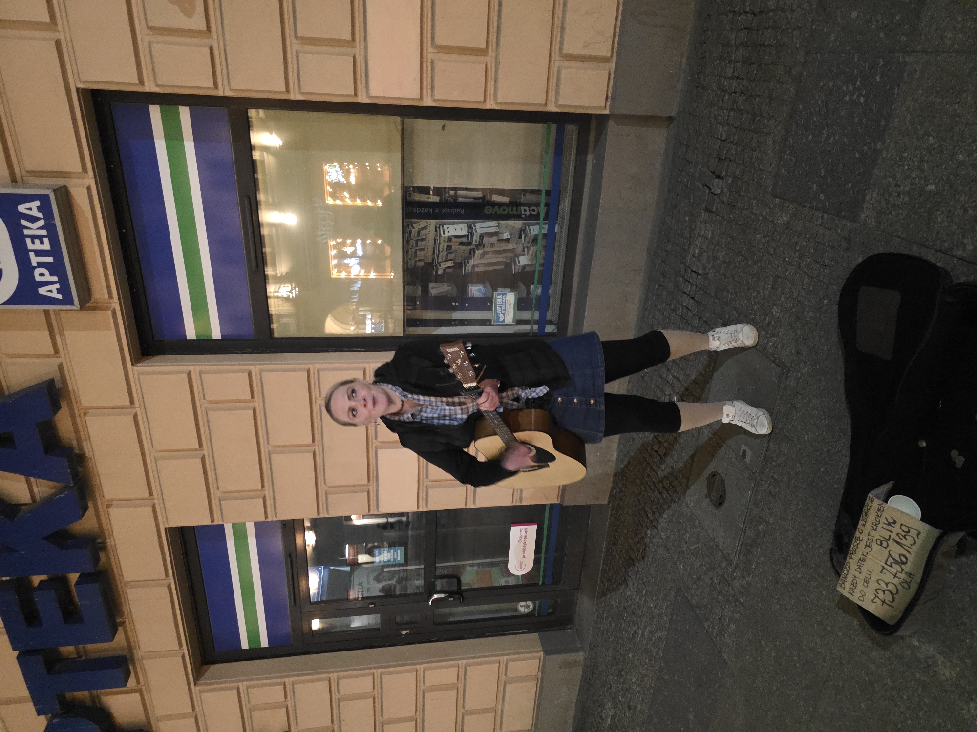 The image captures a street scene at night in what appears to be Warsaw, Poland. A fair-skinned woman with blonde hair is standing on a paved street, playing an acoustic guitar. She is dressed in leggings, sneakers, a denim skirt, a checkered shirt, and a black jacket. The woman is facing the camera, looking toward the lens.
Behind her is a beige, tiled building facade with an "APTEKA" (Pharmacy) sign in blue letters. The pharmacy shopfront features a large window with blue, white, and green vertical stripes. The interior of the pharmacy is partially visible through the reflection and window, with product displays and lighting.
To her right, on the pavement, is a black guitar case and a cardboard sign with handwritten Polish text and a BLIK (mobile payment) number. The sign asks for support, indicating she might be busking.