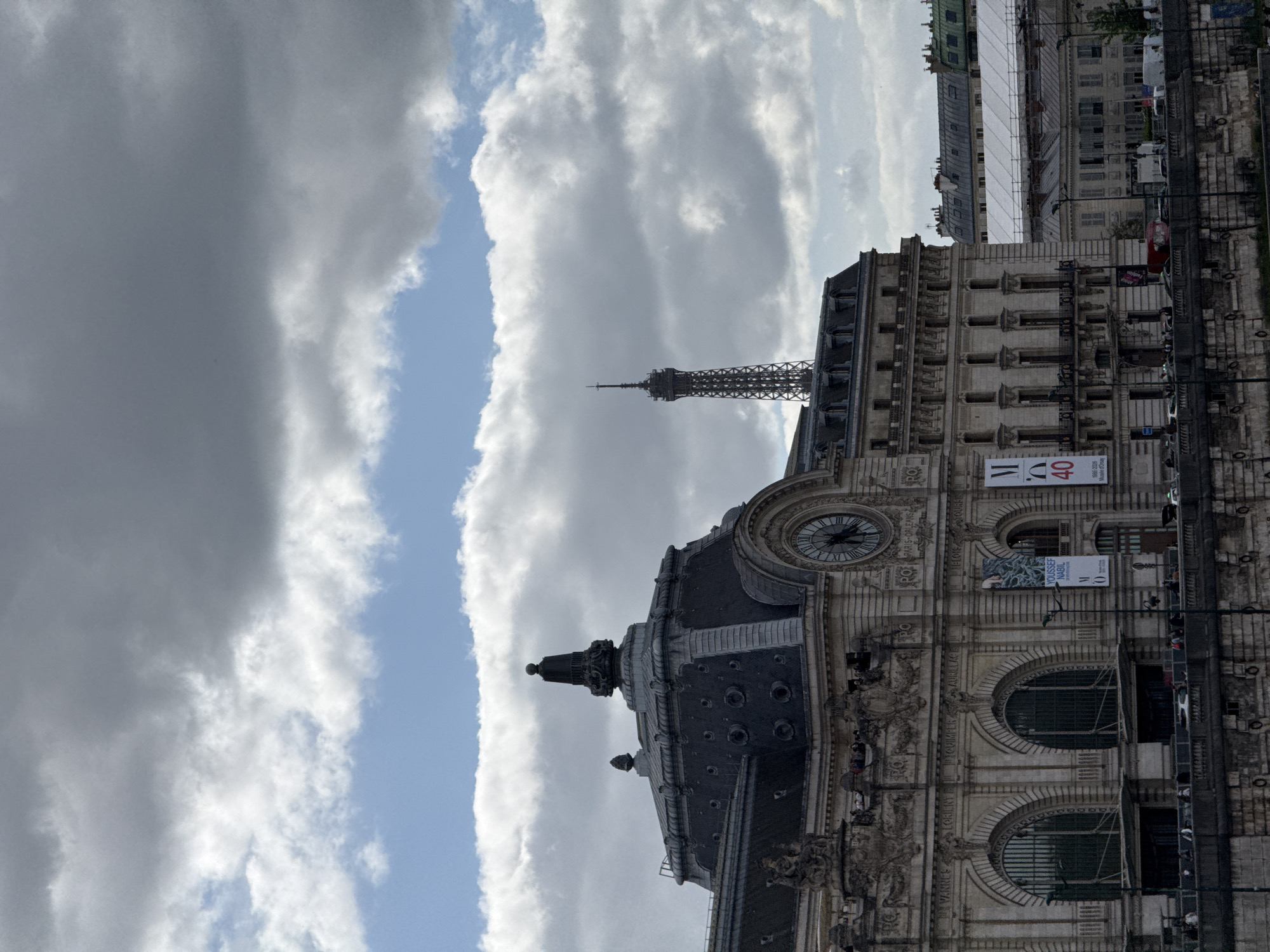 The image captures a vibrant street scene in Paris, centered around a grand, ornate building identified as the Musée d'Orsay. This prominent structure, with its elaborate classical facade, takes up the right side of the frame and features a large, dark-faced clock with Roman numerals near its roofline. Visible banners on the building confirm its identity and feature text such as "M 40" and "1980-2020 Musée d'Orsay," suggesting a 40th-anniversary celebration. Another banner advertises "YOUSSEF NABIL." The building's roof is dark and domed, contrasting with the lighter stone of its walls, which include multiple large arched windows at street level.
In the mid-ground, slightly to the left and rising above the other structures, is the distinct silhouette of the Eiffel Tower, a quintessential Parisian landmark. Beyond the foreground building, other multi-story Parisian buildings can be seen, some featuring scaffolding, indicating ongoing maintenance or construction.
The sky is a dominant element, filled with dramatic, voluminous grey and white clouds. A prominent, narrow band of bright blue sky cuts through the clouds on the far left, creating a striking visual contrast. The scene appears to be set during the daytime, likely an overcast day with intermittent sun peeking through. At the base of the Musée d'Orsay, glimpses of vehicles and pedestrians suggest a bustling urban environment. The image presents a classic view of Paris, blending iconic architecture with a dynamic sky.