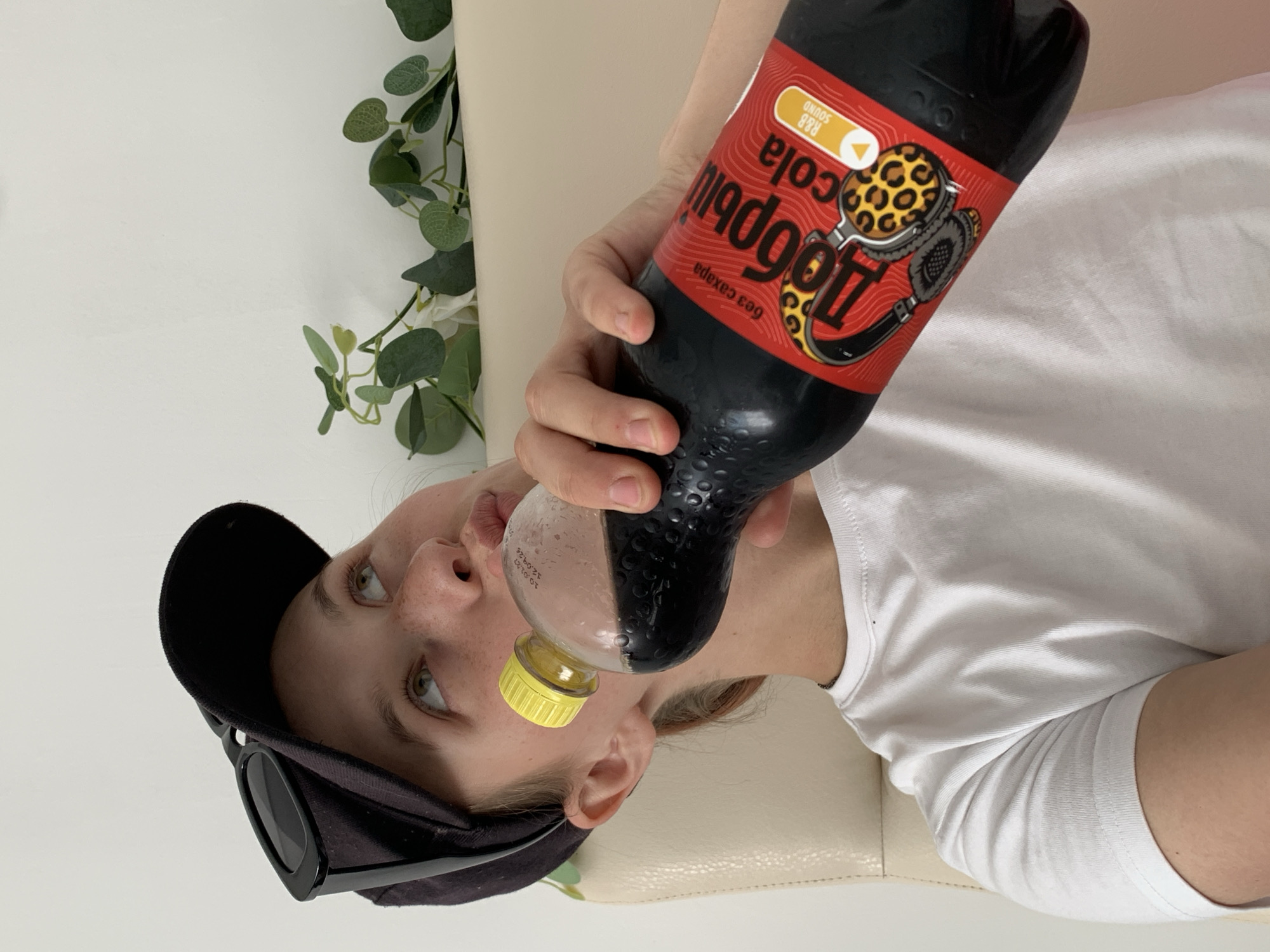 A woman is holding a bottle of "Добрый Cola R&B Sound" beverage with a yellow cap to her mouth as if drinking from it. She is wearing a black baseball cap with sunglasses hanging from the brim. The woman is wearing a white t-shirt and is seated on a light-colored surface. In the background, there is a green plant against a white wall. There is no evidence that this photo was taken in the city of Фоки, Russia.