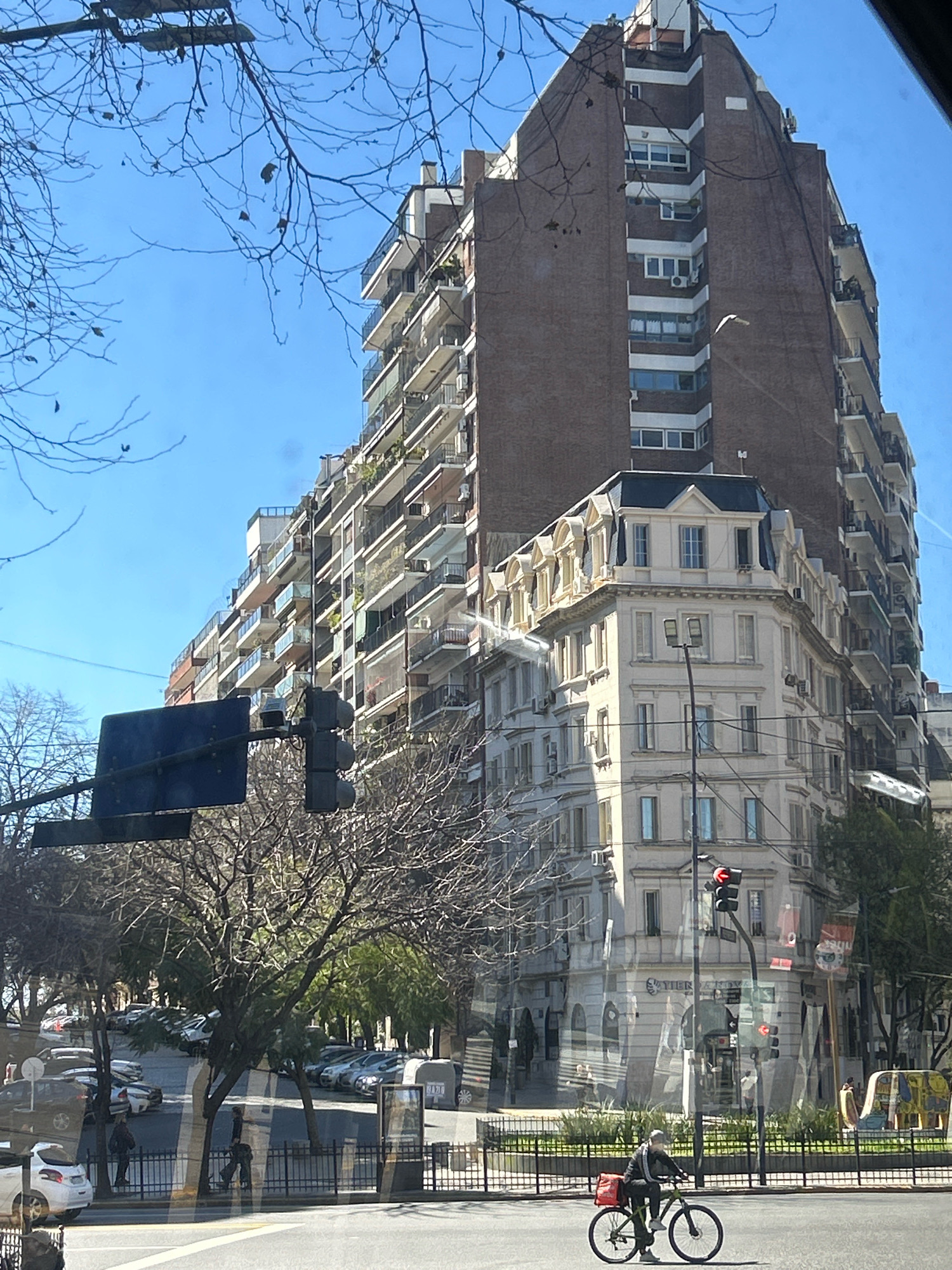 The image depicts a street scene in Buenos Aires, Argentina, under a clear blue sky. In the foreground, a person is riding a bicycle on the right side of the street. The bicycle is dark-colored, and the rider is wearing dark clothing and a cap, with a red bag attached to the back of the bicycle. A traffic light is visible, displaying red, indicating the bicycle rider is stopped at the intersection.
Behind the bicycle, a black metal fence encloses a planter area with sparse vegetation. Beyond the fence, trees with bare branches are prominent, indicating a cooler season. Further in the background, cars are parked along the street and in a lot.
Dominating the background are several buildings. On the right, a tall, multi-story brick building with white accents and numerous balconies stands out. To its left, a cream-colored building with classical architectural features, including dormer windows and ornate detailing, is situated at a corner. Another large building, partially visible on the far right, is also visible. A traffic signal and a dark blue sign are mounted on a pole in the mid-ground. The overall impression is a busy urban environment with distinct architectural styles.
