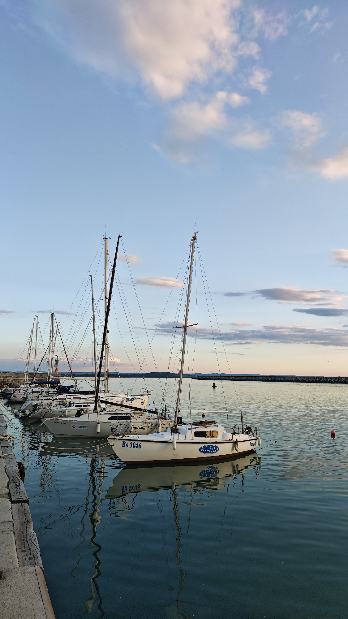 The image depicts a harbor in Burgas, Bulgaria. Several sailboats are docked at a concrete pier. The sailboat closest to the viewer is white with the inscription "Bs 3046" and "hi-lite". A body of water reflects the boats and masts. The sky is a light blue color with white and gray clouds. In the background, there is a stretch of land. The lighting suggests it may be early morning or late afternoon.