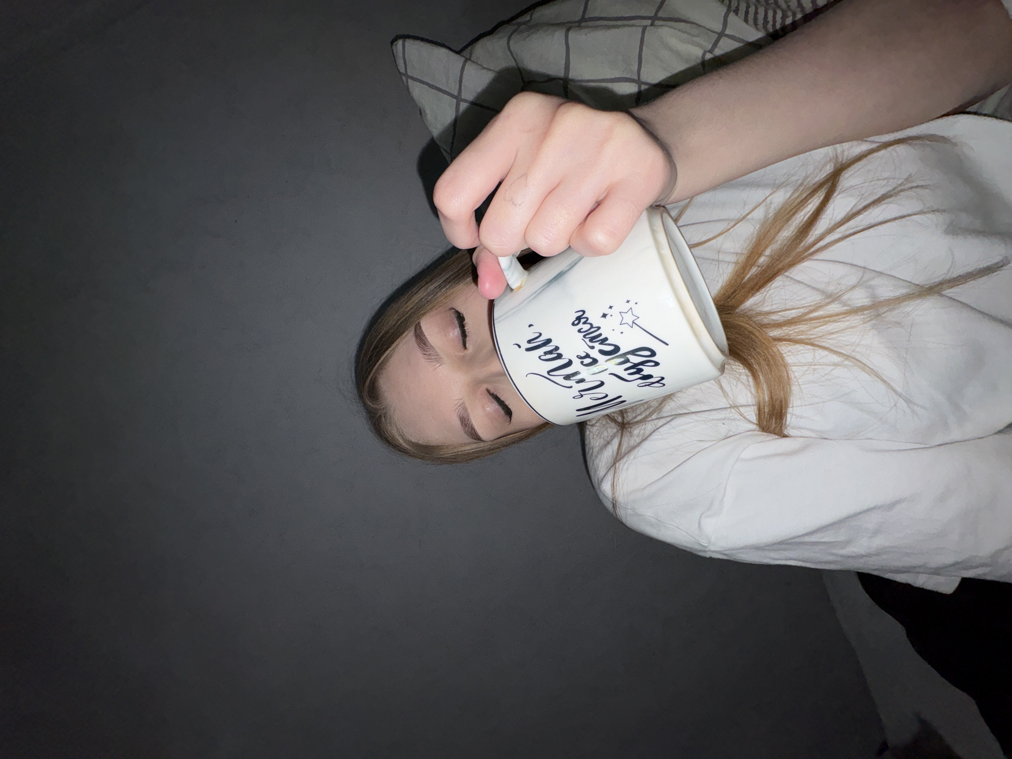 The image presents a slightly overhead, close-up view of a young person in an indoor setting, likely a bedroom in Chaikovsky, Russia. The person, with light-colored hair, lies with eyes closed, possibly asleep or resting. Her face is partially obscured by a white mug held in her right hand, which covers her forehead and eyes. She is wearing a white long-sleeved top. A grey and white checkered pillow is visible behind her head, and the background is a plain dark grey surface.
The activity appears to be a playful or symbolic representation of morning or rest. The person's hair spills out from the mug, suggesting a creative pose. The dim lighting implies it might be early morning, late evening, or a darkened room.
Visible text on the white mug, written in a dark, cursive font, clearly reads "Morning," followed by "magic begins" (or "magic comes") along with a small star icon, adding to the theme of a new day or a morning ritual. The overall mood is gentle and somewhat whimsical.