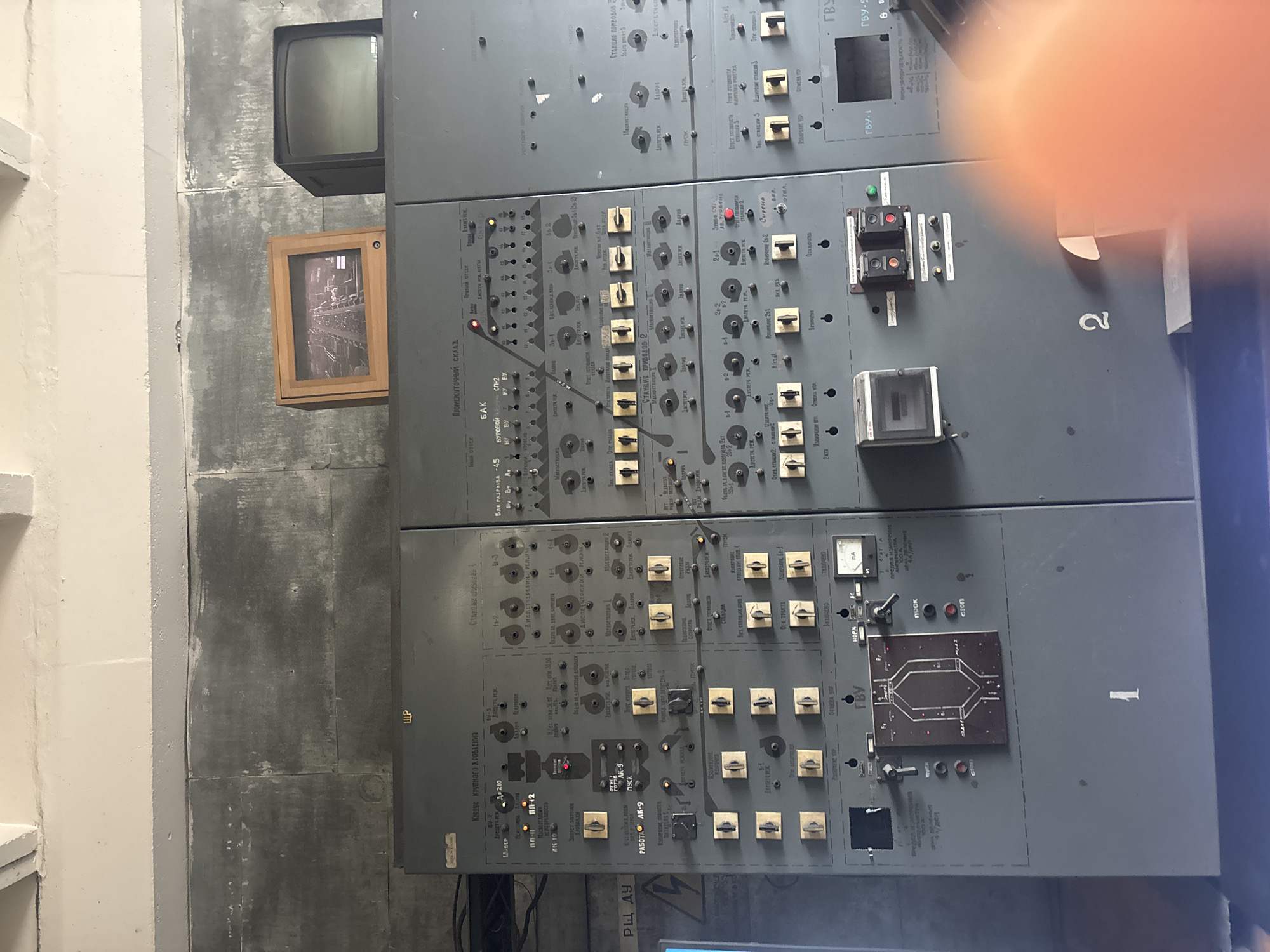 In a dimly lit room with grey concrete walls and a white ceiling, a large, grey industrial control panel dominates the foreground. The panel is covered in numerous buttons, switches, knobs, and displays, many of which are labeled in Cyrillic script. To the left of the panel, a blue surface is visible, possibly a table or another piece of equipment, with a yellow warning sign featuring a lightning bolt.
On top of the control panel, two screens are placed. The one on the left is a smaller, framed digital display showing a grainy, black and white image of what appears to be a railway track or industrial setting. To its right is a larger, older television set with a dark screen.
The control panel itself is divided into sections, each with distinct labels and controls. Some labels indicate functions like "Промежуточный склад" (Intermediate warehouse), "СТАНЦИЯ ПРИБОРОВ" (Instrument station), and "БАК" (Tank). There are also indicators and meters, including a small analog meter labeled "mA" (milliamperes). Various toggle switches and push buttons are present, some with small red or green indicator lights. A prominent diagram of a stylized circuit is located in the lower left section of the panel.
Two large numbers, "1" and "2", are stenciled on the front of the control panel, likely indicating its position or designation. The overall impression is of a complex, potentially dated, technological control system in an industrial or scientific facility. The specific location of Kryvyi Rih, Ukraine, is not directly indicated by any visual elements in the image itself, but this context would be based on external information.