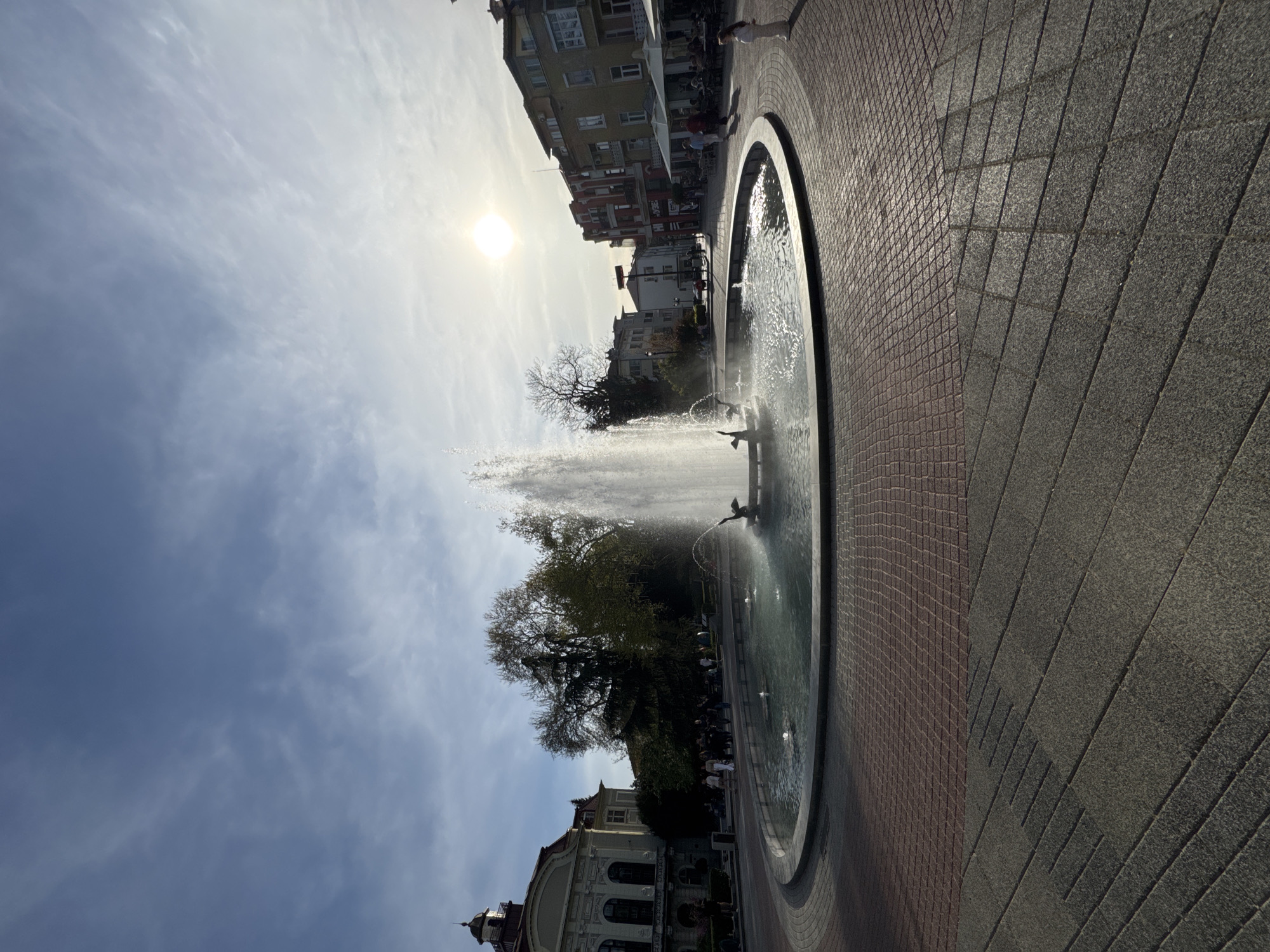 The image shows a water fountain in a public square in Plovdiv, Bulgaria. The fountain is circular with a wide, shallow basin. Water is shooting up from the center of the fountain and from several smaller nozzles around the edge. Buildings are visible in the background, including a domed structure and multi-story apartment buildings. Trees are also visible between the fountain and the buildings. The sky is partly cloudy with the sun shining through. The plaza surrounding the fountain is paved with two different colors of square stones. People are walking near the fountain.