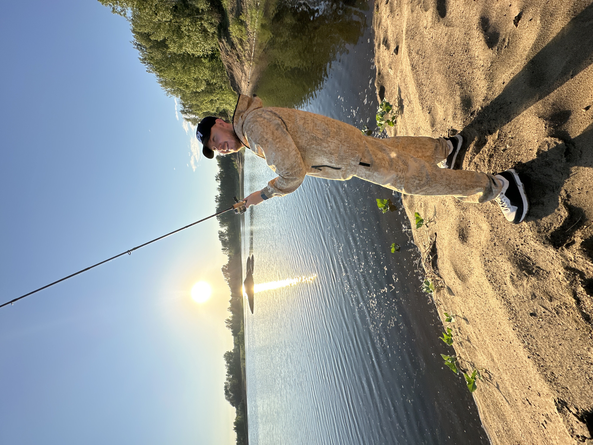 A man is fishing on the bank of the Kama River in Chaikovsky, Russia, during sunset. The sun is low on the horizon, casting a warm golden light on the scene. The man, dressed in camouflage fishing gear and a black baseball cap, holds a fishing rod. The river reflects the sunlight and the surrounding trees, creating a serene atmosphere. The sandy bank is dotted with small green plants.