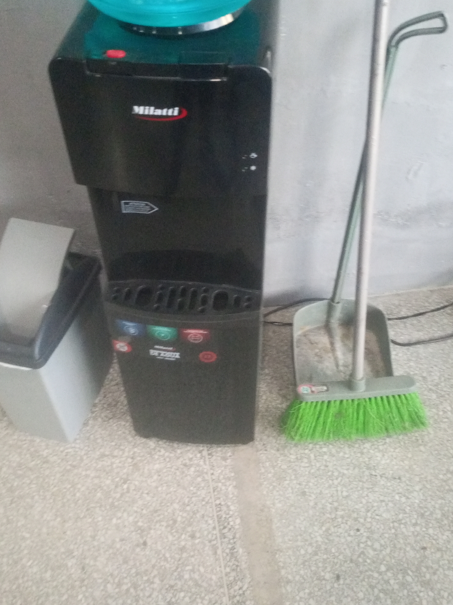 In Juan Griego, Venezuela, a black water dispenser with a turquoise top sits on a mottled gray floor, adjacent to a light gray trash can. To the right of the dispenser, a green broom with a gray handle rests in a gray dustpan, positioned against a textured gray wall. The scene appears to be indoors, possibly in a utility area or a common space. There are no people visible in the image. The lighting suggests an interior environment.