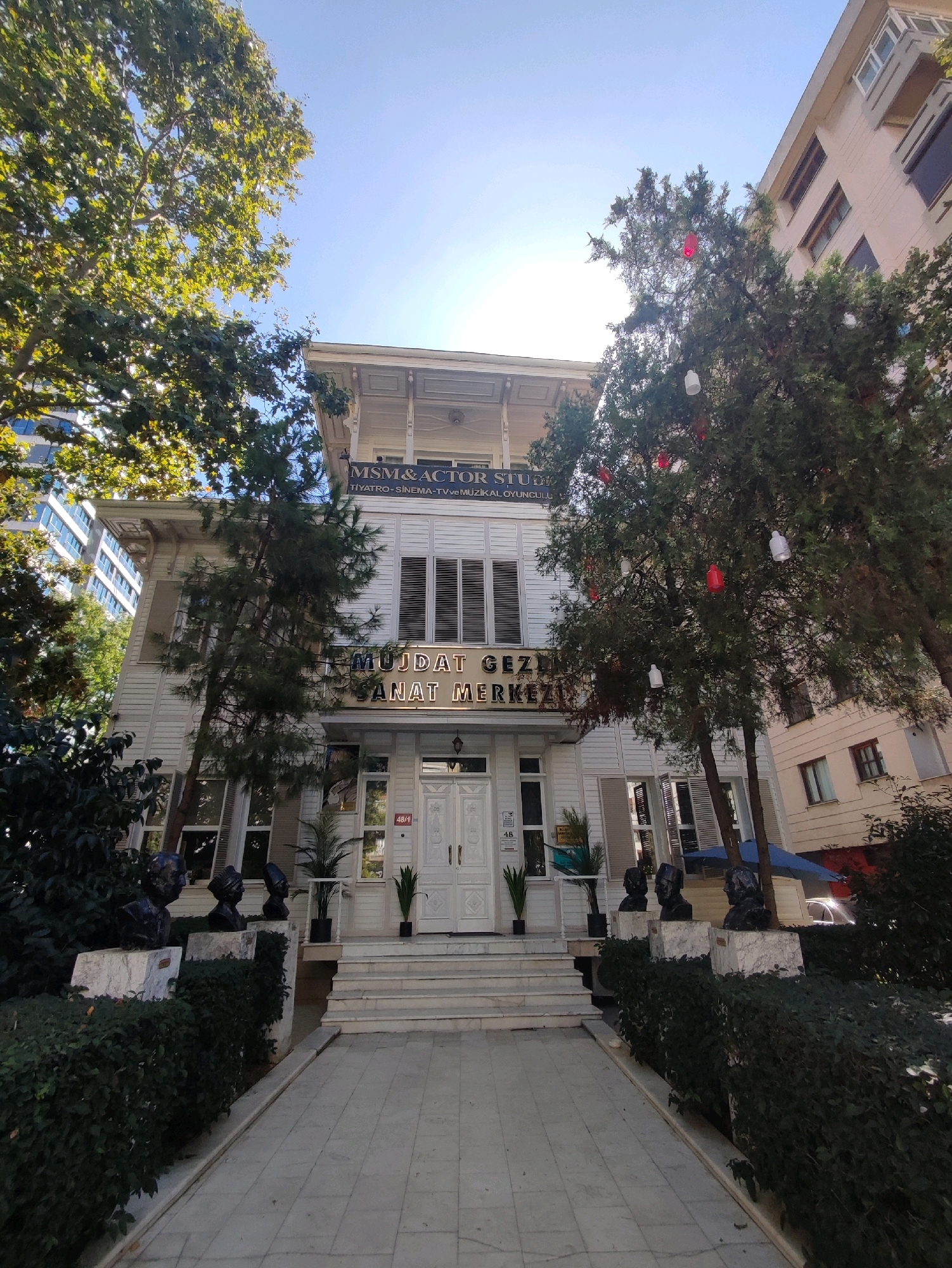 The image captures the facade of a white, two-story building with a prominent entrance. The building features large windows with slatted shutters and decorative architectural details. Above the entrance, a sign reads "MSM & ACTOR STUDIO" and below it, "MUJDAT GEZEN SANAT MERKEZI". The building is surrounded by lush greenery, including trees with decorative lanterns hanging from their branches, and meticulously maintained bushes. Three busts are prominently displayed on white pedestals in front of the bushes, to the left of the entrance. To the right of the entrance, a modern apartment building is visible. The scene is bathed in sunlight, suggesting it is daytime. The location is identified as Mujdat Gezen Sanat Merkezi in Kadıköy, Turkey, a cultural center. There are no people visible in the image.