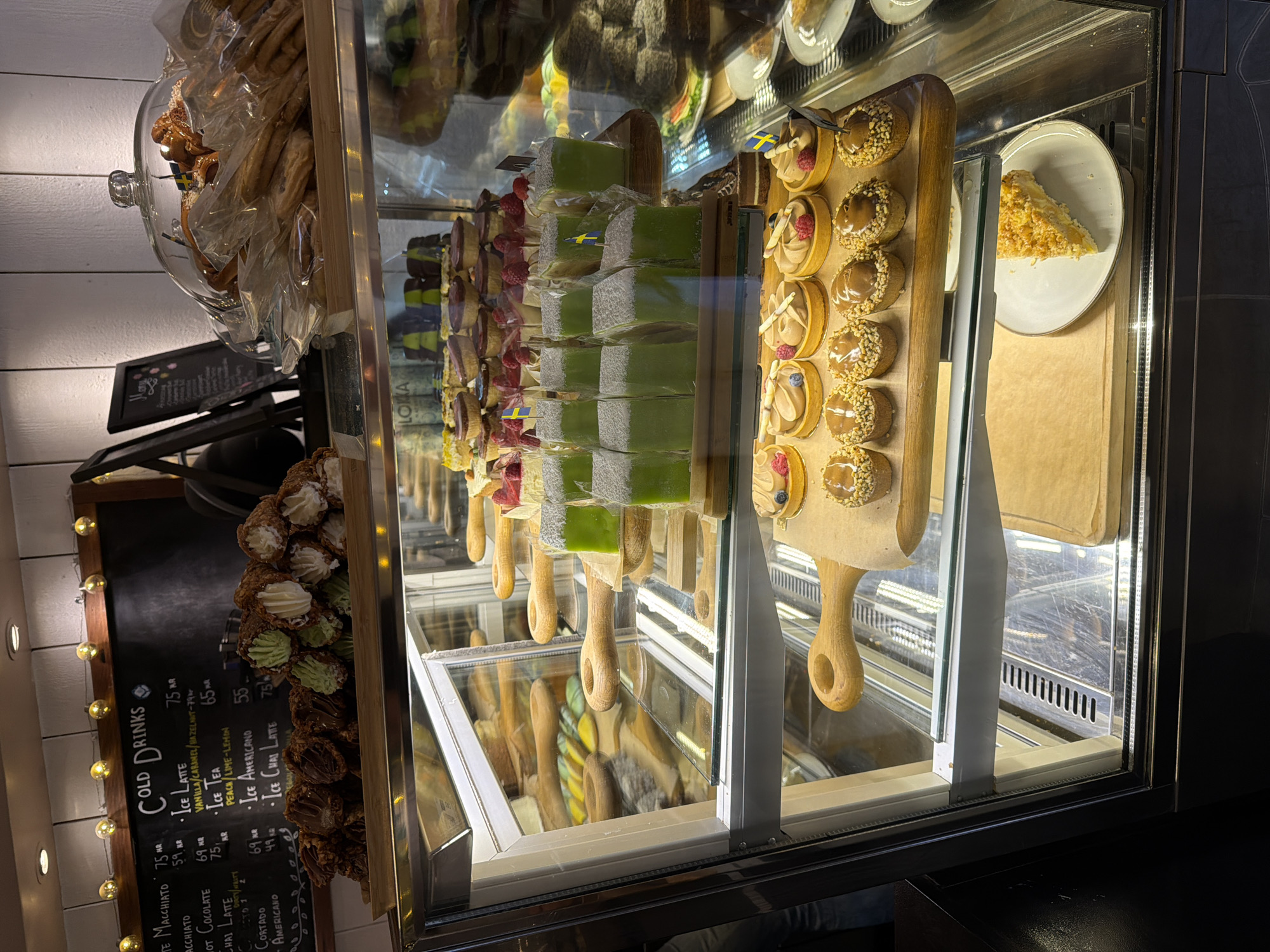 The image depicts the interior of a bakery or cafe, likely located in Stockholm, Sweden, given the presence of Swedish flags on some pastries and the context of the menu board. The scene is a close-up view of a refrigerated display case filled with a variety of baked goods and desserts.
In the upper section of the display case, several rows of small green cakes, possibly Prinsesstårta or a similar confection, are visible, each topped with a miniature Swedish flag. These cakes are arranged on a wooden board. To the left of these cakes are several chocolate-covered pastries and what appear to be ice cream cones filled with cream and a green filling.
Above the display case, a chalkboard menu lists "COLD DRINKS" with various coffee and tea options like "Ice Latte," "Ice Tea," and "Ice Americano," along with their prices in Swedish Krona (kr). String lights are hung above the chalkboard.
Further down in the display case, more desserts are showcased, including tarts with fruit and cream toppings, and smaller, individual pastries with nuts and caramel. A single slice of what appears to be apple pie or a similar fruit tart is presented on a white plate on a lower shelf.
The overall environment suggests a retail setting with a focus on pastries and beverages. The lighting is artificial, illuminating the food items within the display. There are no people clearly visible interacting with the items. The style of the pastries and the menu information point towards a Swedish establishment.
