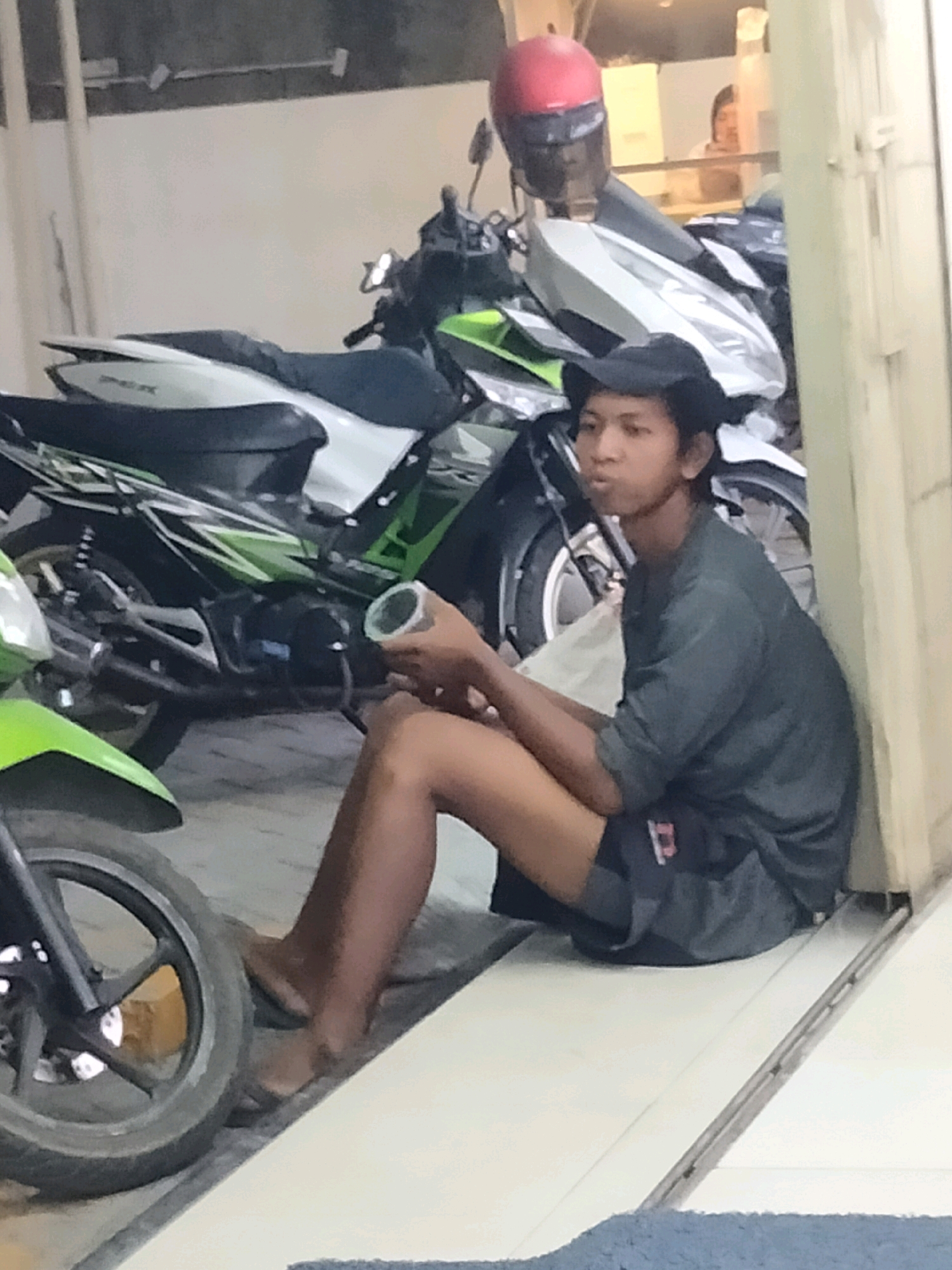 In Pisangan Jaya, Indonesia, a young man wearing a dark t-shirt, dark shorts, and a black baseball cap sits on the floor next to two motorcycles. The man is holding a silver cylindrical object in his right hand. One motorcycle, predominantly white and green, is parked behind him, and another partially visible motorcycle, green and black, is to his left. The environment appears to be an outdoor area, possibly a parking lot or a street, with a light-colored wall and door frame visible to the right. A red helmet rests on the seat of the motorcycle behind the man.