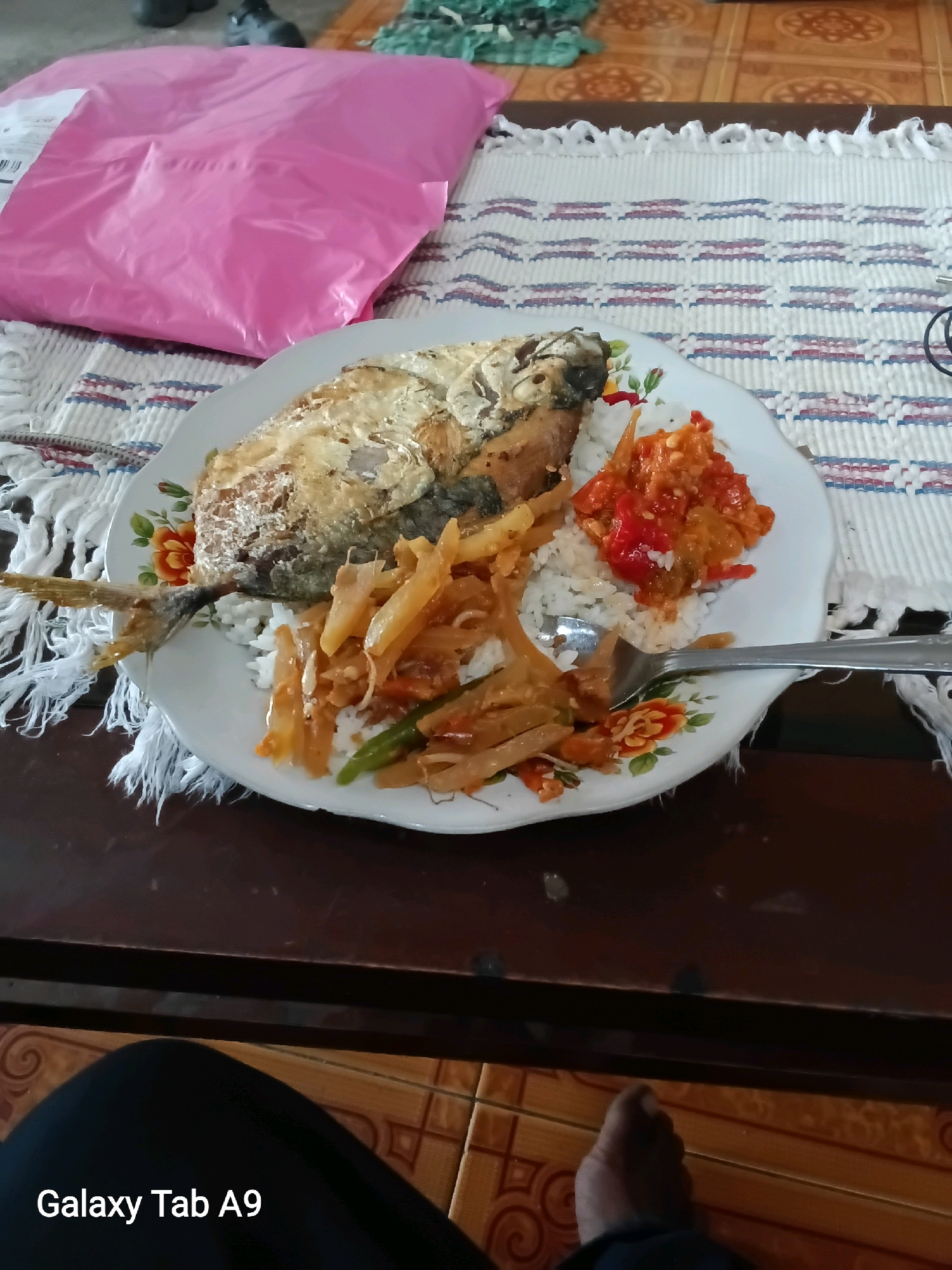 A plate of food is on a table. The plate contains rice, fried fish, and a side dish. A fork is on the plate. In the background, a pink package is on a woven mat. The table is made of wood and the floor is tiled. The text "Galaxy Tab A9" is visible in the lower left corner of the image, indicating that the image was taken with a Samsung Galaxy Tab A9 device. The food appears to be Indonesian cuisine, common in Trenggalek, Indonesia.