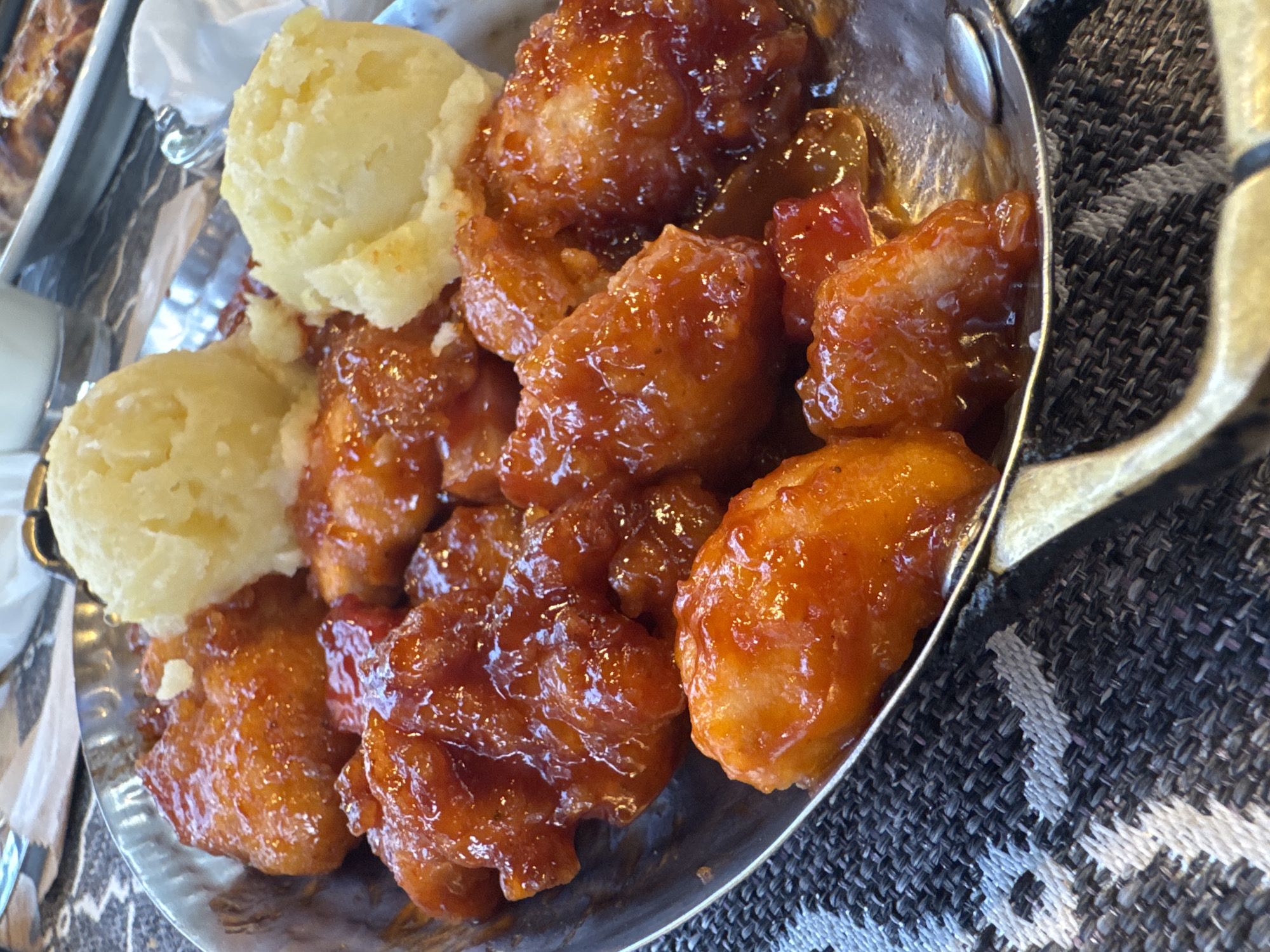 In a close-up, eye-level shot, a metallic bowl filled with glazed food and mashed potatoes is presented. The bowl, with a textured rim, rests on a dark grey and white patterned tablecloth. The food consists of several pieces of what appears to be fried chicken, coated in a glossy, orange-red sauce. Three scoops of pale yellow mashed potatoes are arranged in the bowl, interspersed with the chicken. The background is blurred, but suggests a restaurant setting with a white napkin and part of another dish visible. The lighting is bright, highlighting the sheen of the sauce and the texture of the mashed potatoes. This scene is likely from a restaurant in Primorsko, Bulgaria.