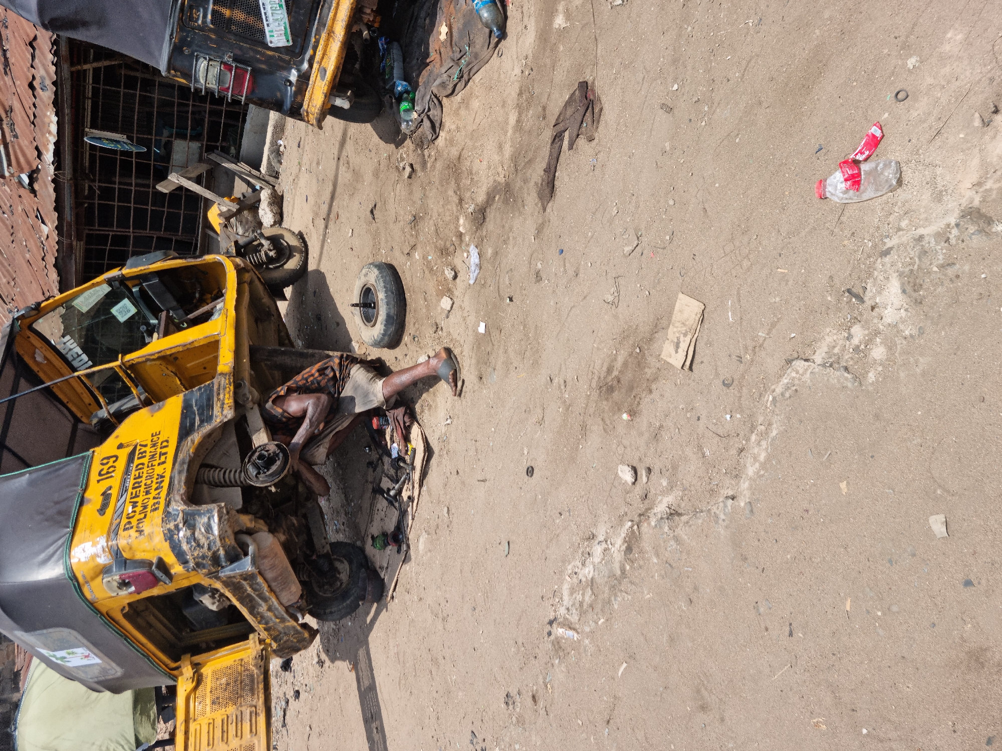 The image captures an outdoor scene in bright daylight, likely in Onitsha, Nigeria, showing a person repairing a yellow keke Napep (auto-rickshaw). The primary subject is a male individual, dressed in patterned shorts and a dark shirt, lying on the dusty, sandy ground to work on the side of the yellow tricycle. One of the keke's wheels is removed and rests nearby, indicating a repair or maintenance task.
In the foreground and midground, two yellow keke Napeps are visible. The one being repaired is prominently featured, with its side panel open and components exposed. Tools and rags are scattered around the work area, along with empty plastic bottles. Another yellow keke Napep is partially visible in the background, also appearing somewhat worn.
The ground is unpaved, consisting of dirt and sand, with scattered debris including pieces of cardboard and plastic. In the background, a building with corrugated metal roofing and a gridded security gate or window is visible, suggesting an urban or semi-urban setting.
Visible text on the side of the keke Napep being worked on includes the number "169" and the phrase "POWERED BY WOLINO MICRO FINANCE BANK LTD". On the license plate of the keke Napep in the background, "LAGOS" and parts of an alphanumeric code "NG-47MB" are discernible. The overall impression is one of a busy, functional roadside repair area under a clear, sunny sky.