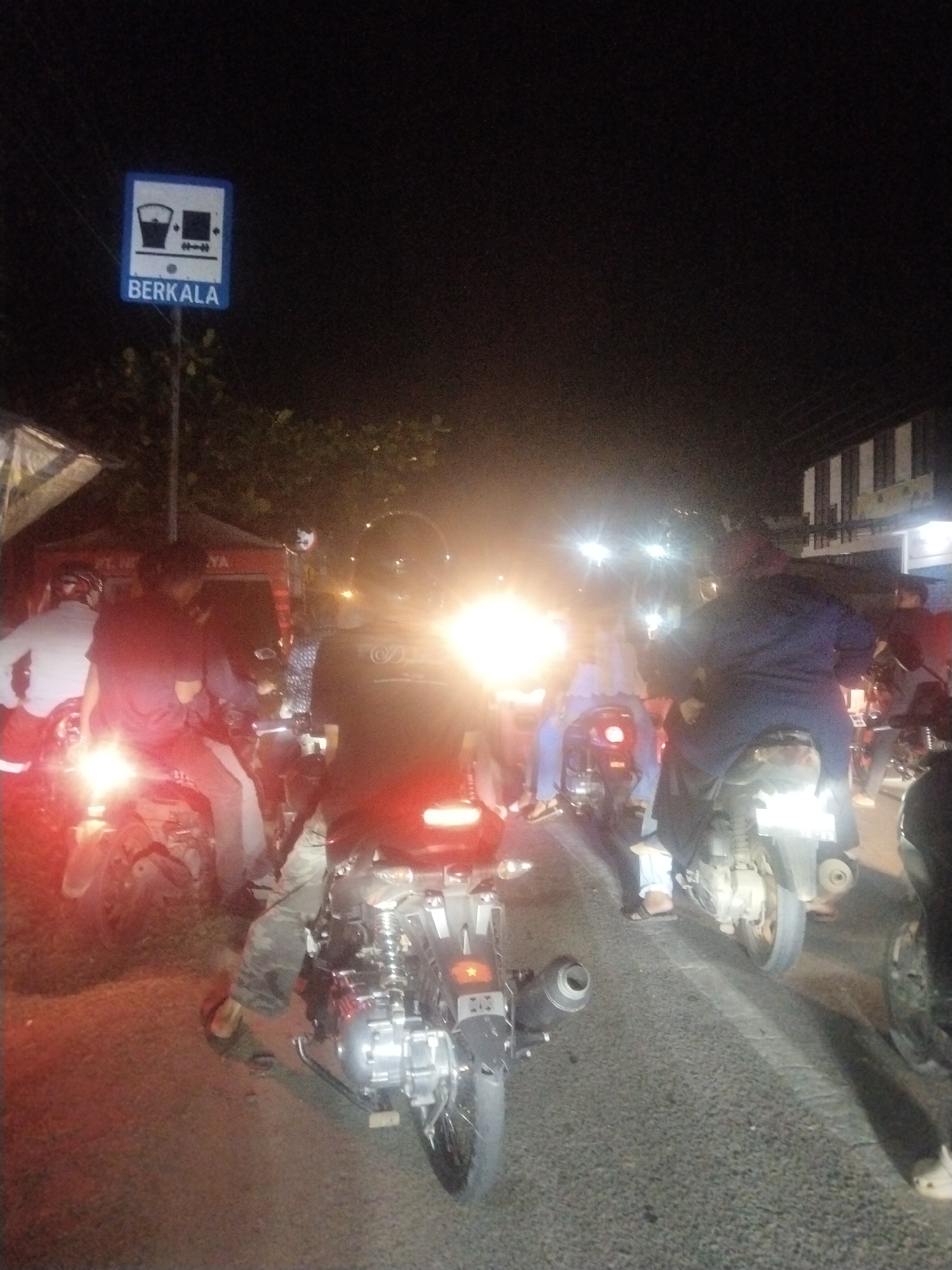 The image shows a group of people on motorcycles at night in what appears to be Lhokseumawe, Indonesia. A blue sign that reads "BERKALA" with symbols depicting a bucket and box is visible on the left side of the image. The motorcycles are mostly stationary, lined up on a road. People are seated on the motorcycles, but their faces are obscured by helmets or darkness. The scene is illuminated by the motorcycles' headlights and taillights, making it difficult to discern details in the background. Buildings are vaguely visible in the background.