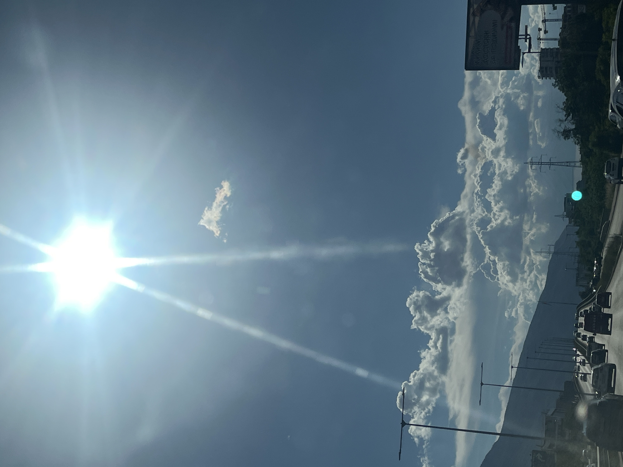 A wide-angle, low-angle shot depicts a bright, midday sun casting strong rays across a clear blue sky. Two prominent sunbeams cut through the atmosphere, originating from the upper left portion of the frame. A single, small, fluffy white cloud is visible in the mid-sky. Below, a multi-lane highway stretches into the distance, filled with a moderate amount of traffic including cars, vans, and trucks. On the left side of the highway, a steep, dark mountainside is partially visible, with streetlights lining the road. On the right side, a billboard with text is present, along with construction cranes and buildings in the distance. The scene appears to be a highway commute within a city.