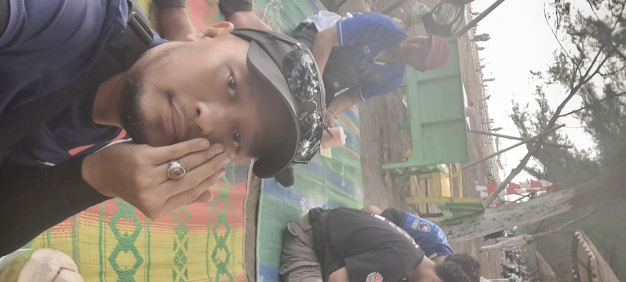 In Nyamplung Sari, Indonesia, a selfie is taken outdoors near a beach. The foreground features a man with a beard and mustache, wearing a dark blue shirt, a black arm sleeve, and a black baseball cap with sunglasses perched on top. He is touching his cheek with his right hand, which is adorned with a ring. Behind him, another man wearing a blue shirt and a red cap is visible, sitting on a mat. Further back, a third person in a black shirt is also seated. They are all on a colorful woven mat on the sand. In the background, there are trees, some beachgoers, and structures that appear to be seating or vendor stalls. The sky is bright, suggesting it is daytime.