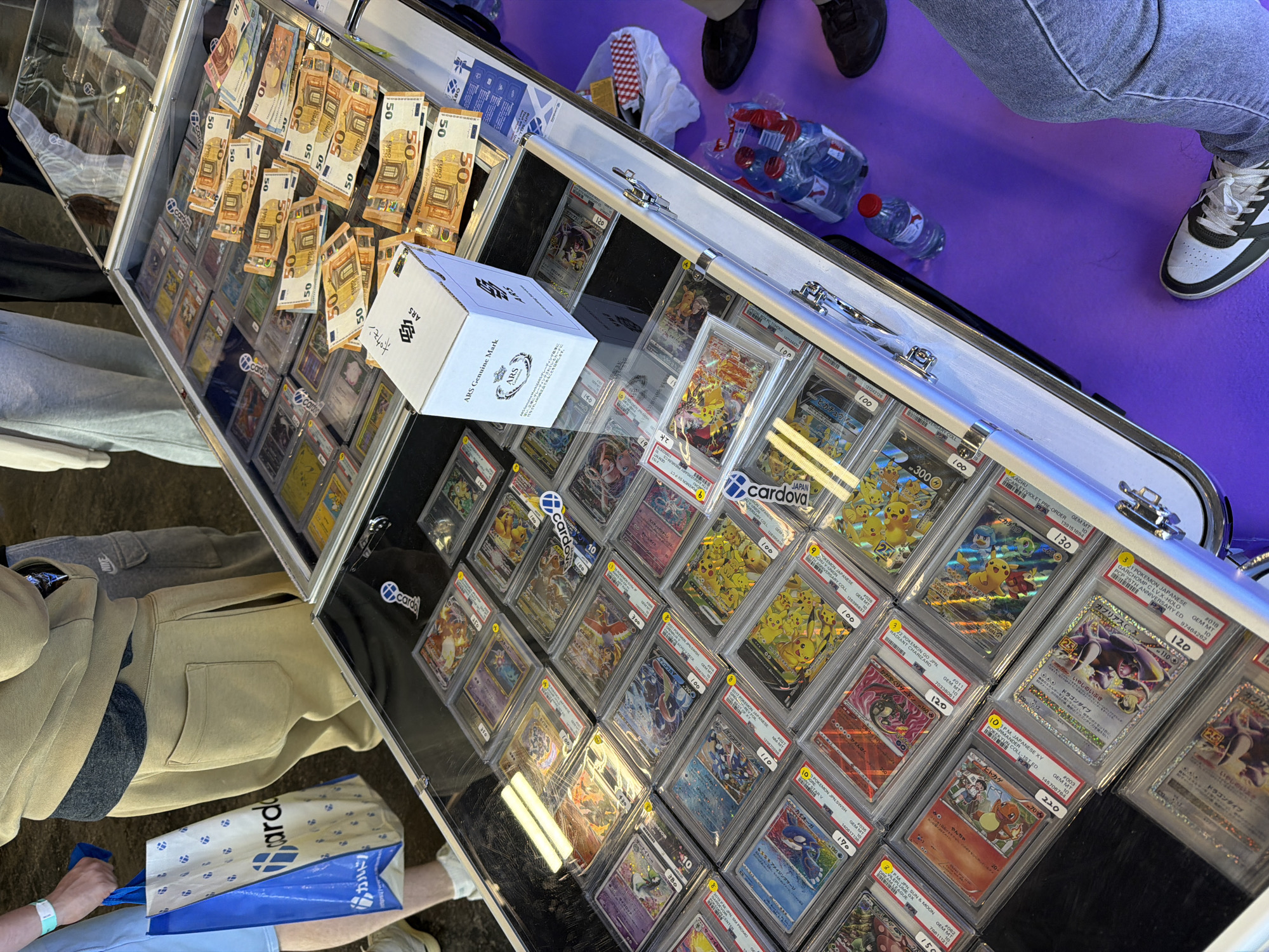 A top-down shot shows a display case filled with graded Pokemon cards, with stacks of Euro bills, mostly 50s, placed on top. A white box labeled "ARS Genuine Mark" sits prominently within the display. The cards are individually cased and priced, with many featuring Japanese text. Surrounding the display are people; one wearing jeans and sneakers, another in khaki pants and holding a blue bag. Scattered around the display case on a purple surface are water bottles and a white bag.