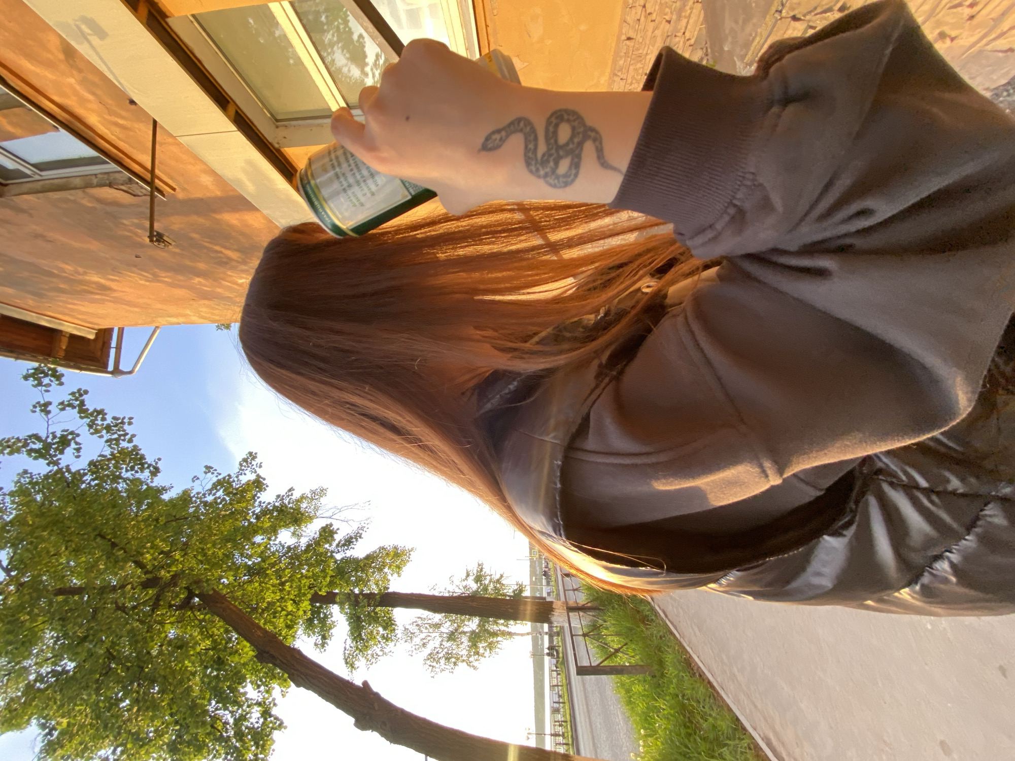 A medium shot, taken from the subject's point of view, shows a woman holding a green beverage can towards the camera, with a snake tattoo visible on her left wrist. She is wearing a dark grey sweatshirt and a puffy black vest. The background features a partially visible building with windows on the upper left, a green tree against a blue sky on the lower left, and a road lined with trees and a body of water on the lower right. The scene appears to be a park or roadside location in Votkinsk, Russia.