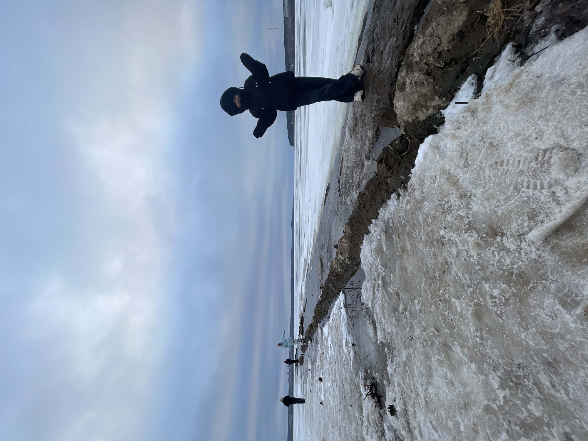 Here is a description of the image:
The image is oriented sideways. It shows a person dressed in dark clothing and white shoes standing on a muddy shoreline covered in ice. In the background, there's a frozen body of water and the cloudy sky. Several people can be seen standing on the ice in the distance. Based on the location, this appears to be a scene in Izhevsk, Russia.