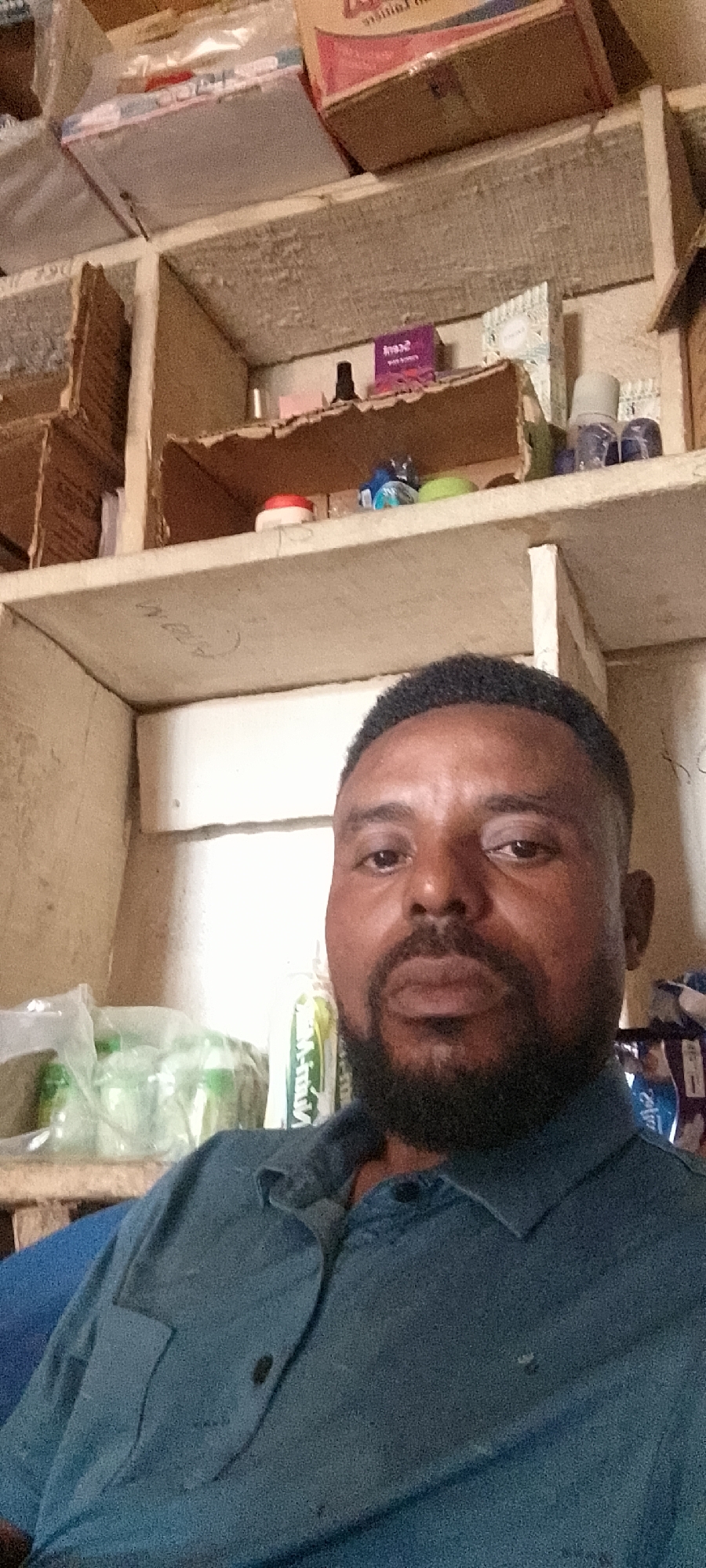 A medium shot, selfie-style image shows a Black man in his 30s or 40s, with dark skin, a dark complexion, and short black hair, facing the camera. He has a full, dark beard and is wearing a dark blue collared shirt. The background suggests a storage or shelf-filled area, possibly in a market or shop in Durumi, Nigeria. Several cardboard boxes and various items are visible on shelves behind him, including plastic bottles, bags, and what appears to be packaged goods. The lighting is somewhat dim, casting shadows and highlighting the texture of the man's skin and beard. The focus is on the man's face and upper torso.