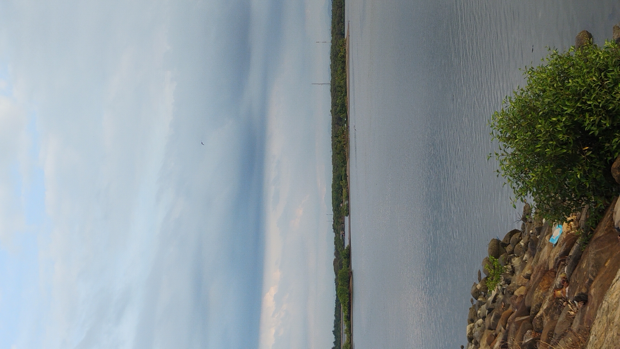 The image depicts a serene natural landscape with a body of water in the foreground and middle ground, likely the Straits of Malacca or a similar coastal area in Lhokseumawe, Indonesia. The water is calm, reflecting the overcast sky. The shoreline is lined with rocks, some of which are partially submerged in the water. Vegetation, including a bushy green plant, is visible on the right side of the shore. In the distance, a strip of land with dense green trees and some man-made structures, possibly communication towers, stretches across the horizon. The sky is filled with clouds, with patches of lighter blue indicating diffused sunlight. A single bird is visible in flight against the sky. The overall scene conveys a sense of tranquility and natural beauty.