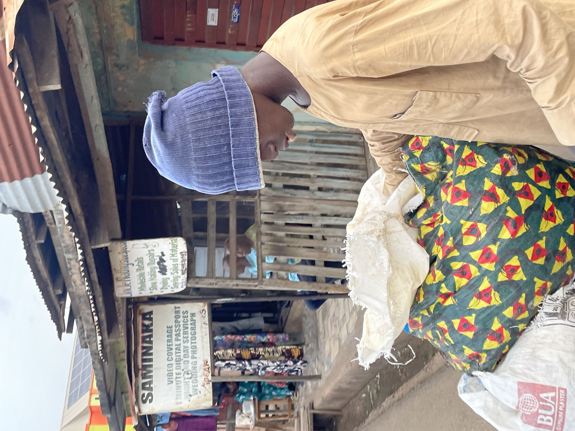 A street scene in an unidentified location, showing a person wearing a blue knit cap and a light brown jacket, leaning over a collection of bags. One bag has a distinctive pattern of yellow and red kite-like shapes on a green background. Another bag has the words "BUA GYPSUM PLASTER" printed on it. In the background, signs advertise "SAMINAKA VIDEO COVERAGE" and "Shoe Making Repairs" along with other services. There are fabric samples or bolts of fabric on display in a shop, as well as other people visible further in the background. The surroundings appear somewhat weathered, with corrugated metal roofing and exposed wooden structures.